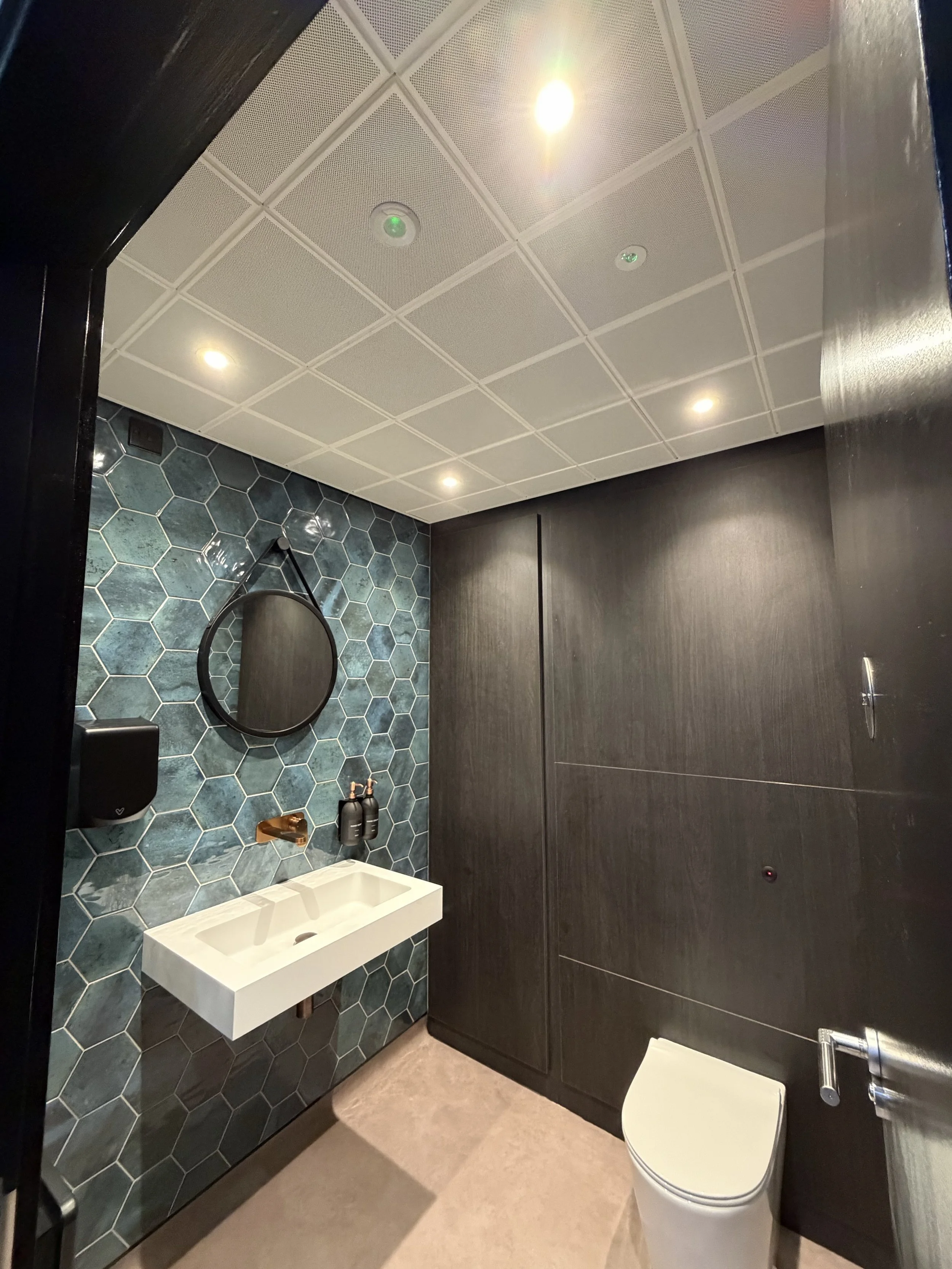 Refurbished washroom at Portman Finance, Brackmills, with ceiling downlights, teal hexagonal tiles, a basin and toilet.