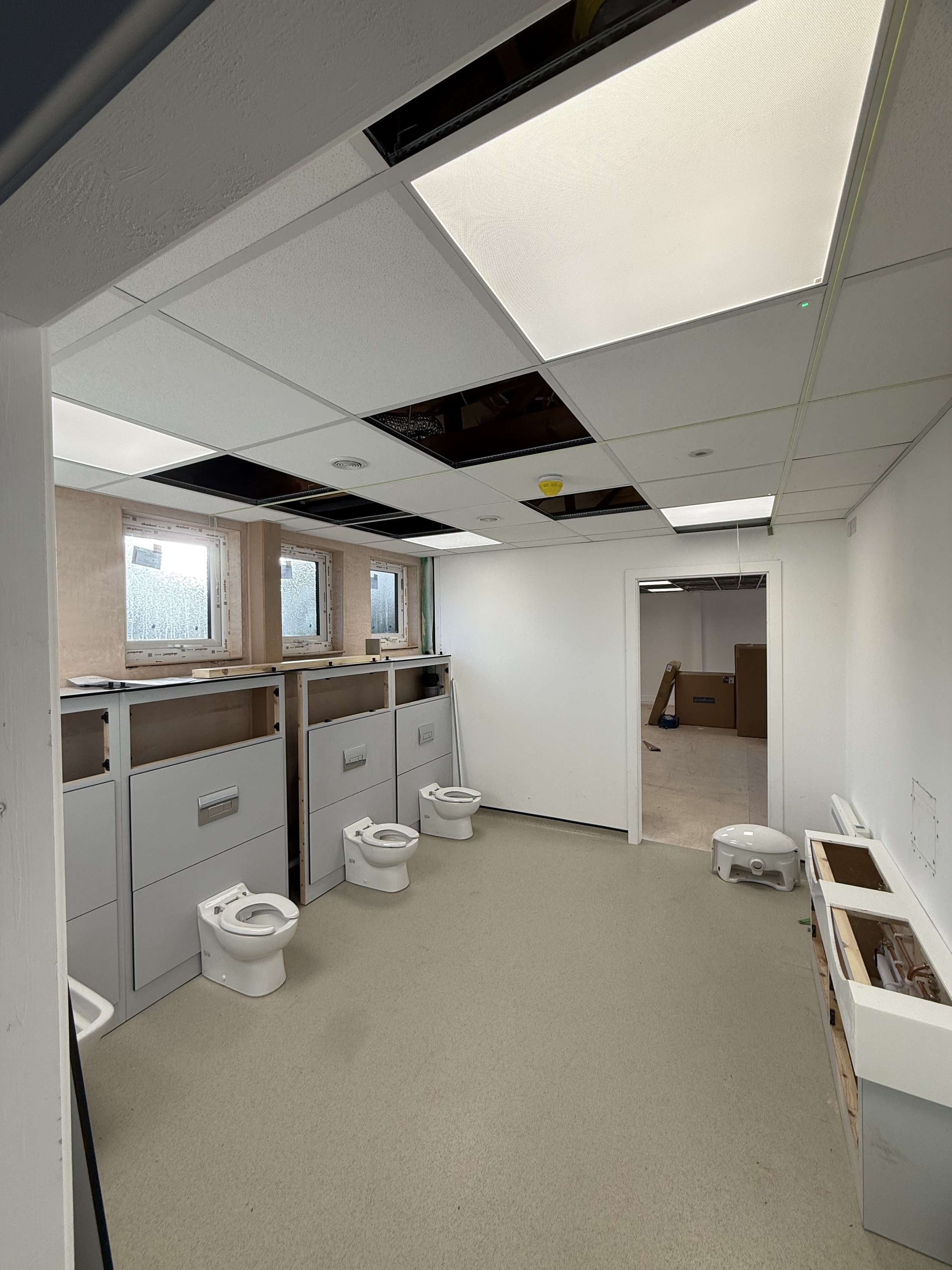 An unfinished bathroom with three toilets, three small windows, and a suspended ceiling with some panels missing.