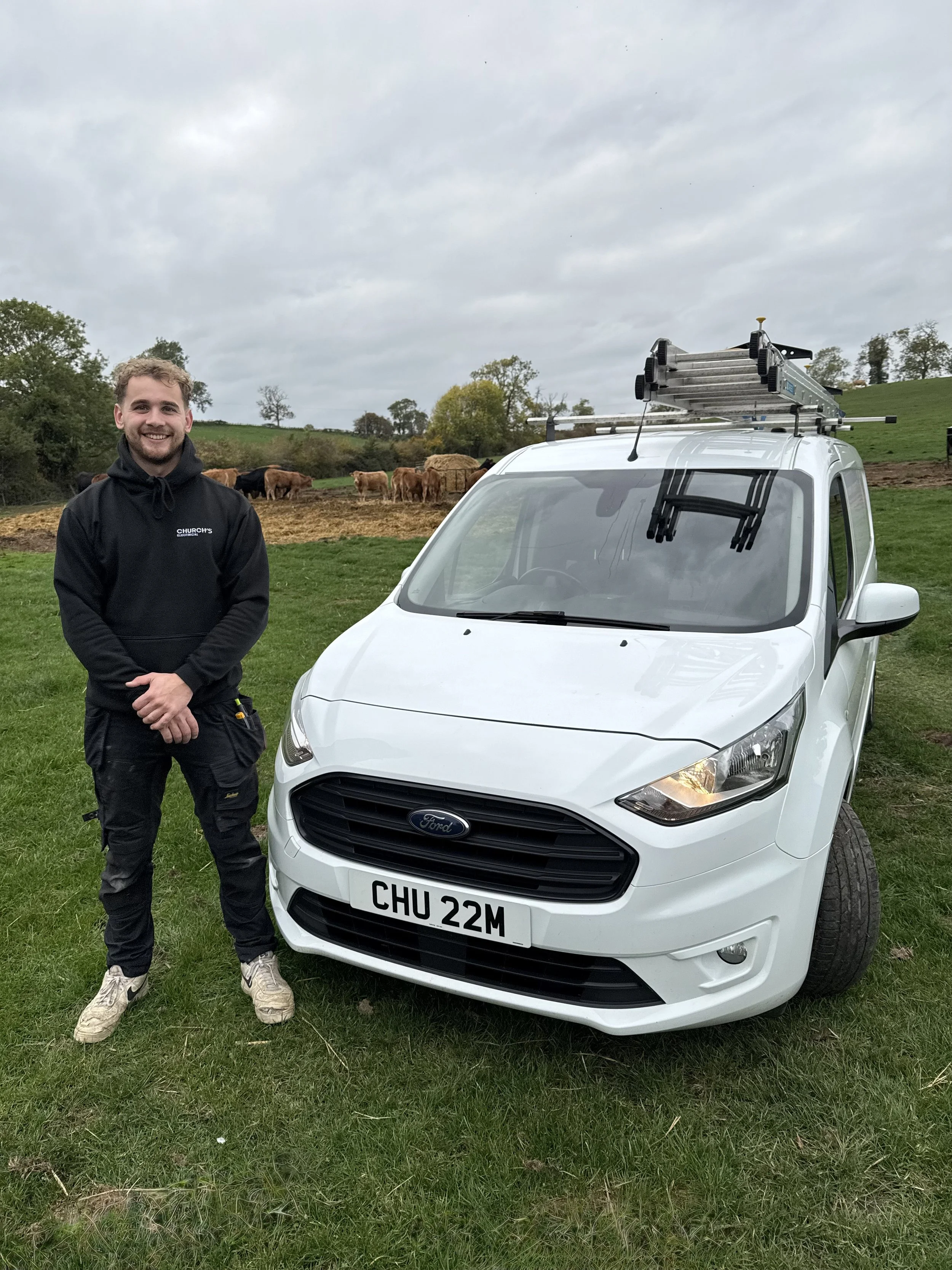 A young man with a black hoodie and black pants standing next to a white Ford vehicle with a roof rack, on a grassy field with cows grazing in the background under an overcast sky.
