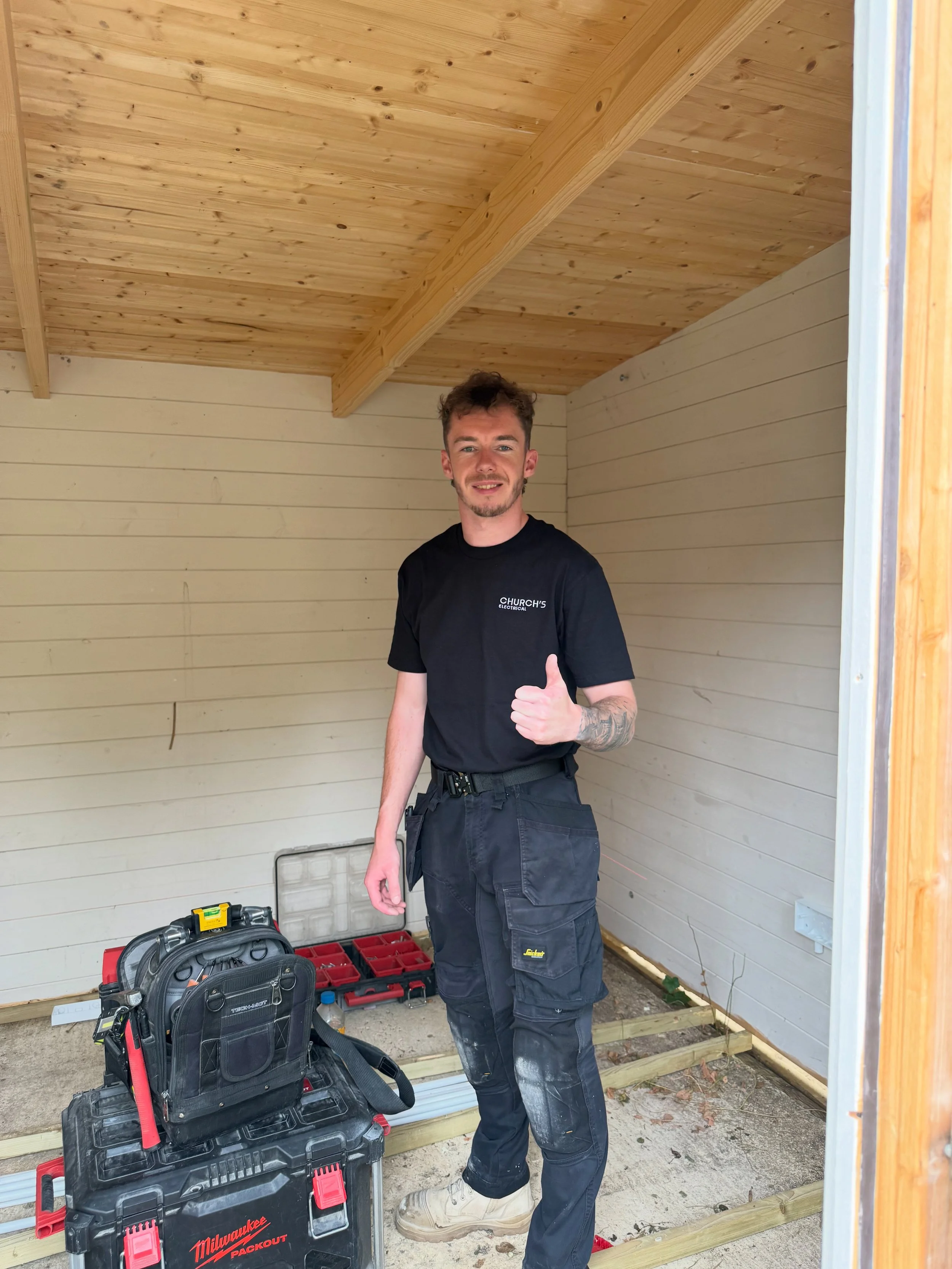 A young man standing inside a partially built wooden structure, giving a thumbs up and smiling, with construction tools and materials around him.