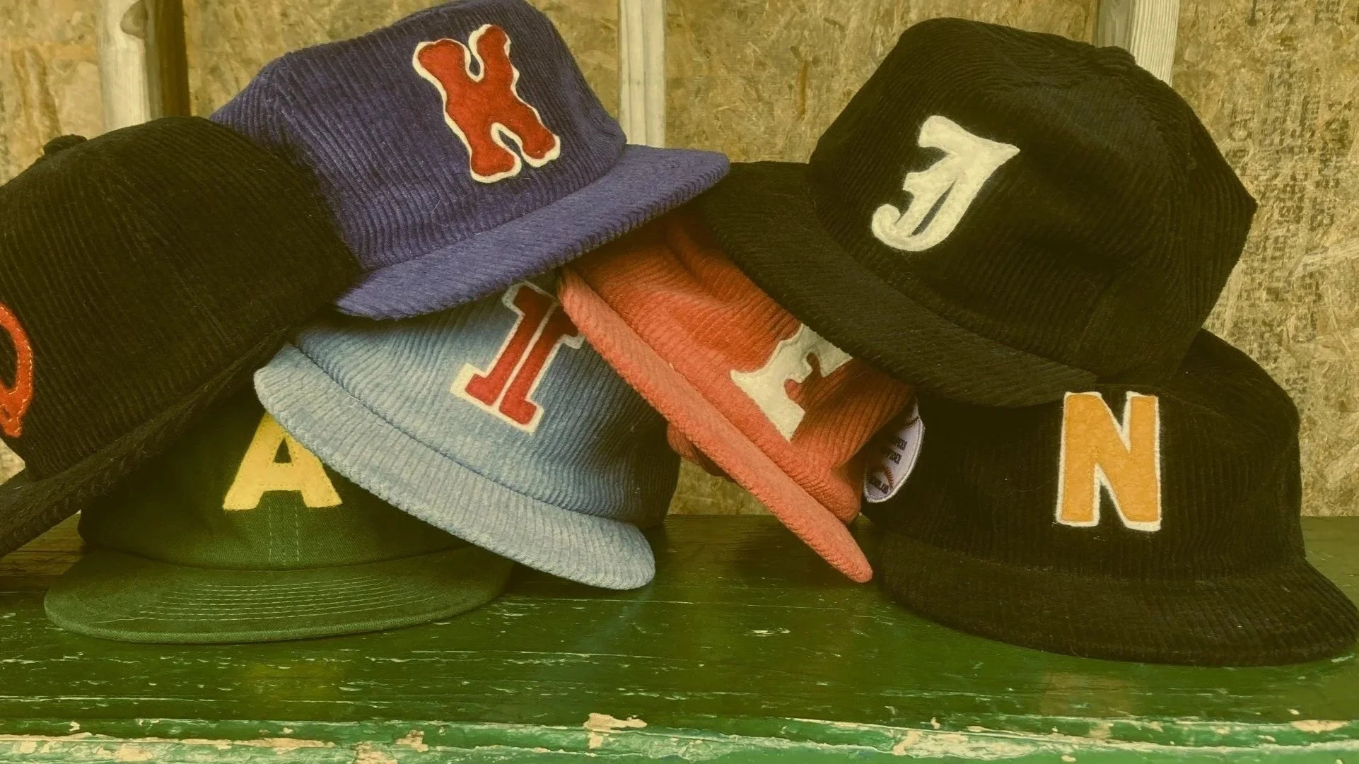 A collection of baseball caps with various colored embroidery of letters and logos, stacked on a green wooden surface.