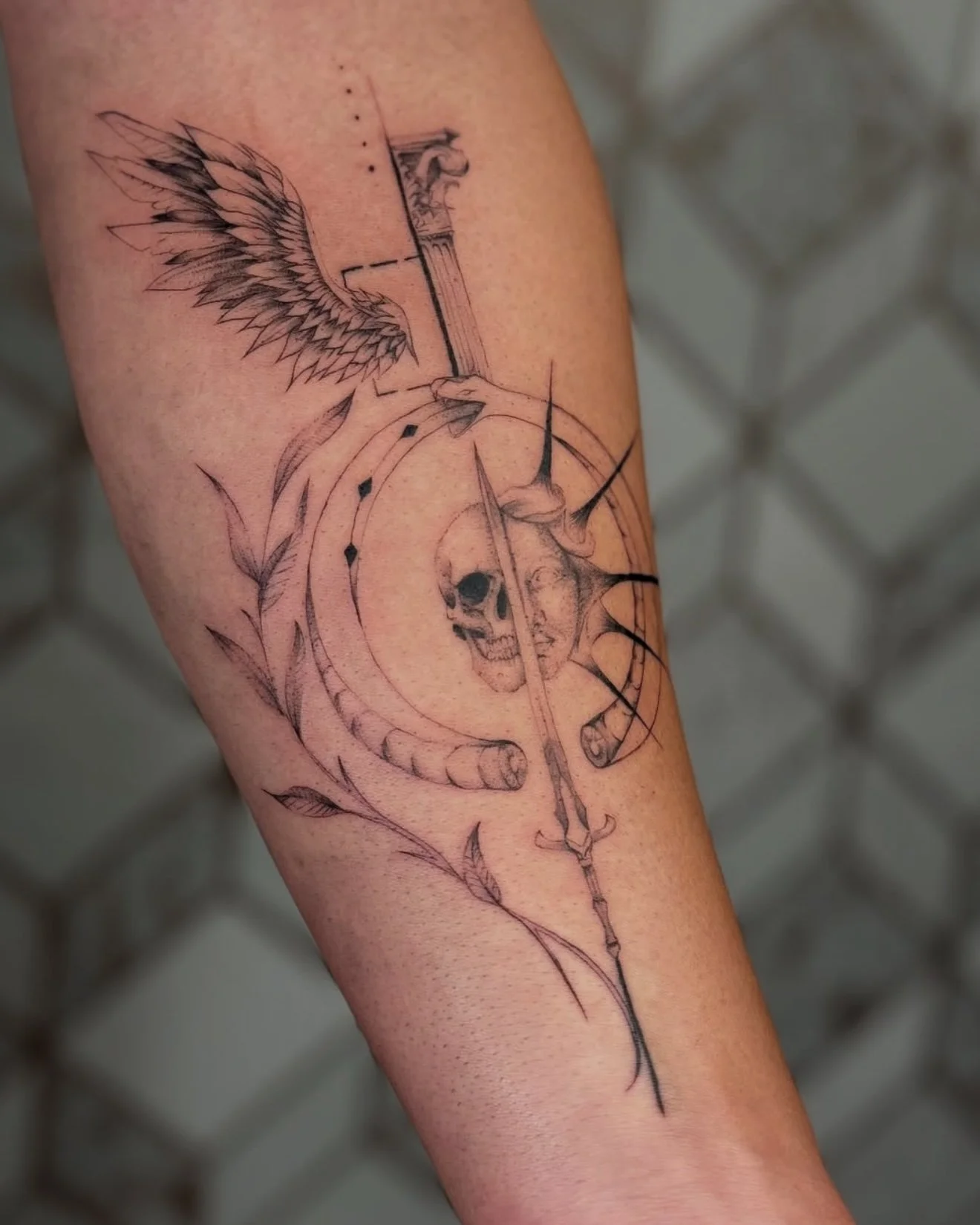 Tattoo of a wing, skull, sword, and Latin text on a person's arm, with various decorative elements.