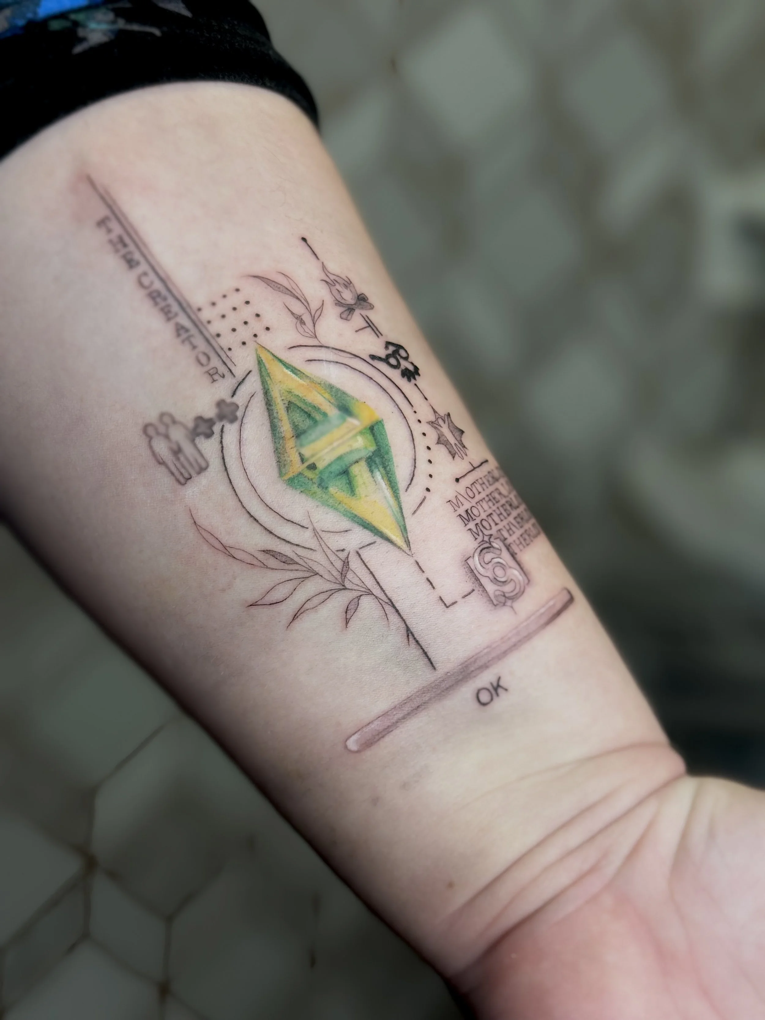 Tattoo on person's arm featuring a colorful geometric diamond shape at the center, surrounded by various small sketches, lines, and words like 'mother' and 'ok'.