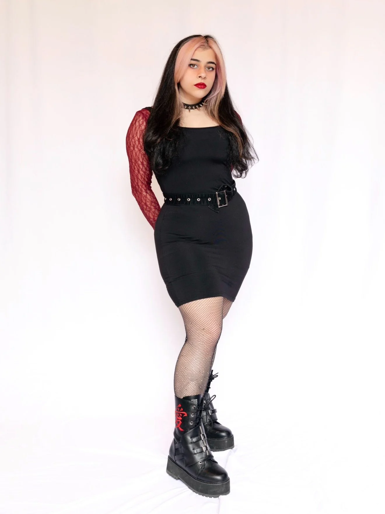 A young woman stands against a plain white background. She has long dark hair with pink streaks, wearing a black dress with red lace long sleeves, a studded choker, a belt, fishnet stockings, and black platform boots with red writing.