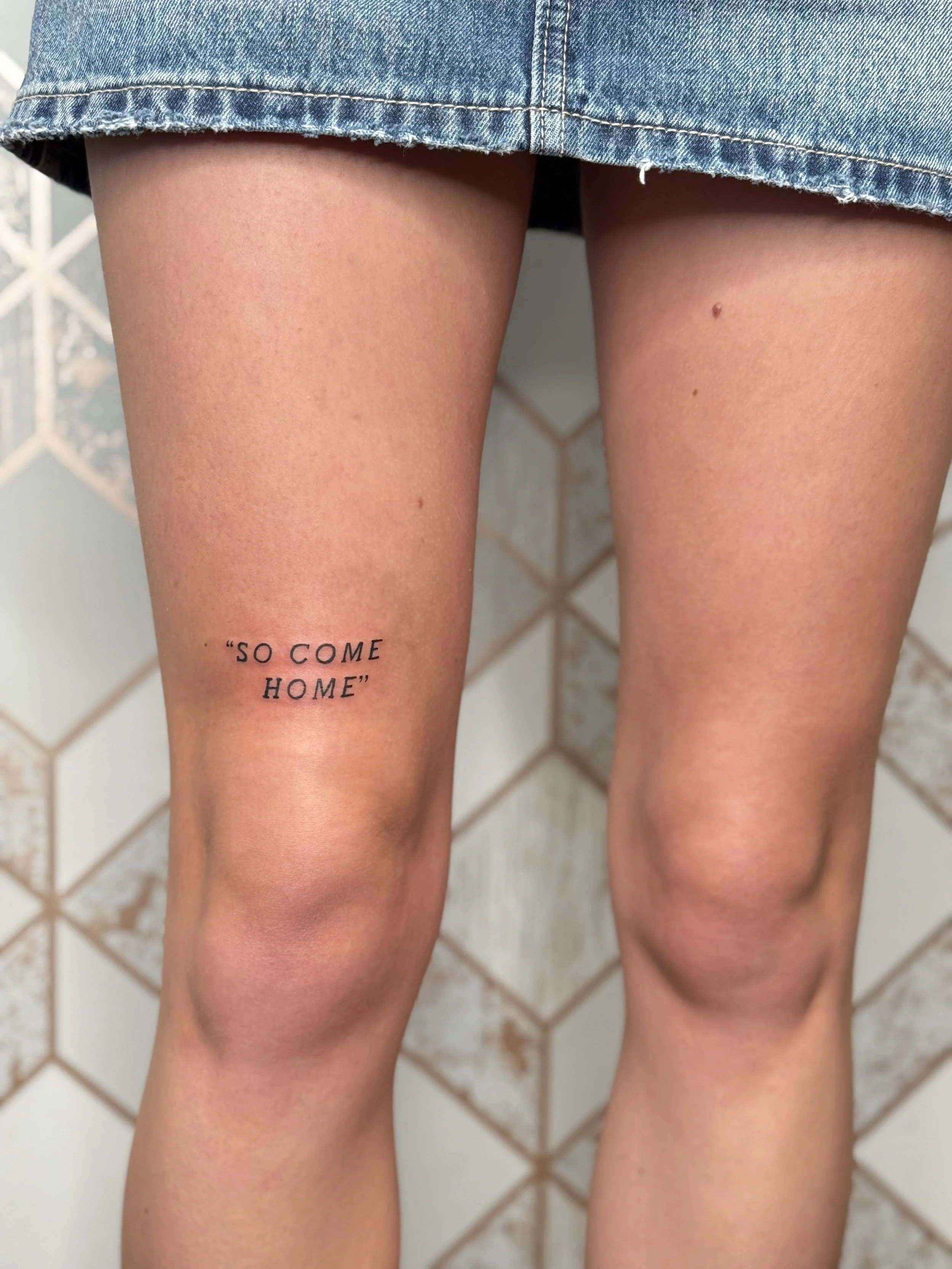 Fine line script quote tattoo reading “so come home” placed above the knee