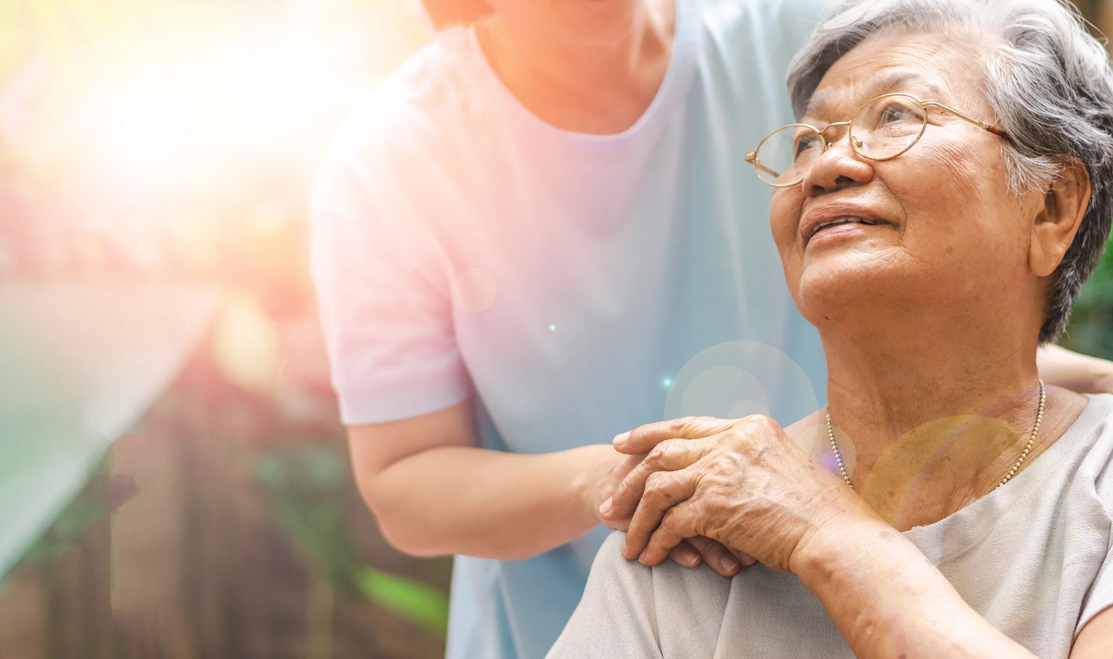 An elderly woman wearing glasses sitting outdoors with sunlight, being embraced by a caregiver which symbolize elder abuse and estate litigation which are issues addressed by Holian Law, P.C., a California Family law firm.