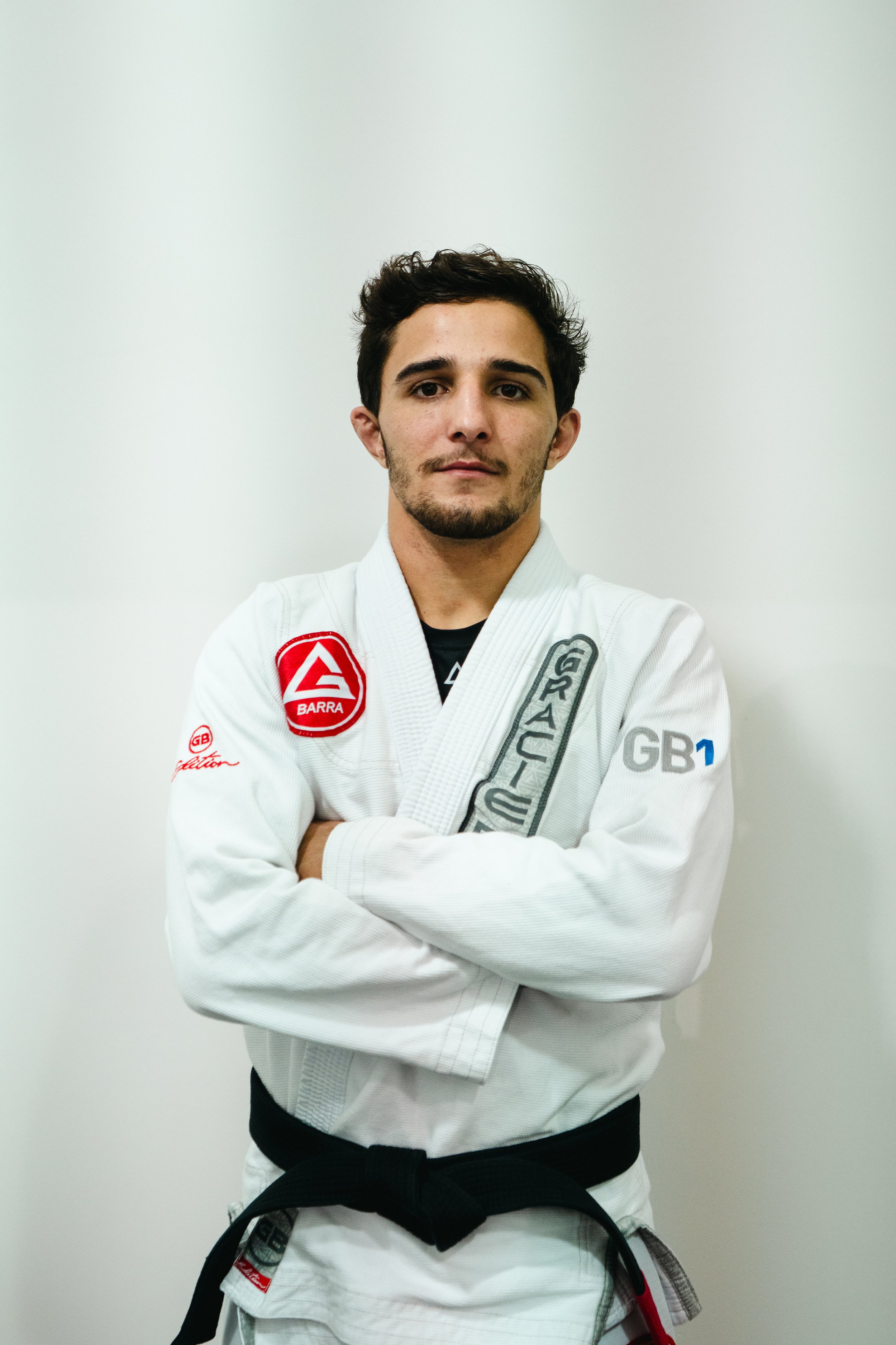 Xandy of Gracie Barra Bryan wearing a white Brazilian Jiu-Jitsu gi with patches, standing with arms crossed against a plain background.