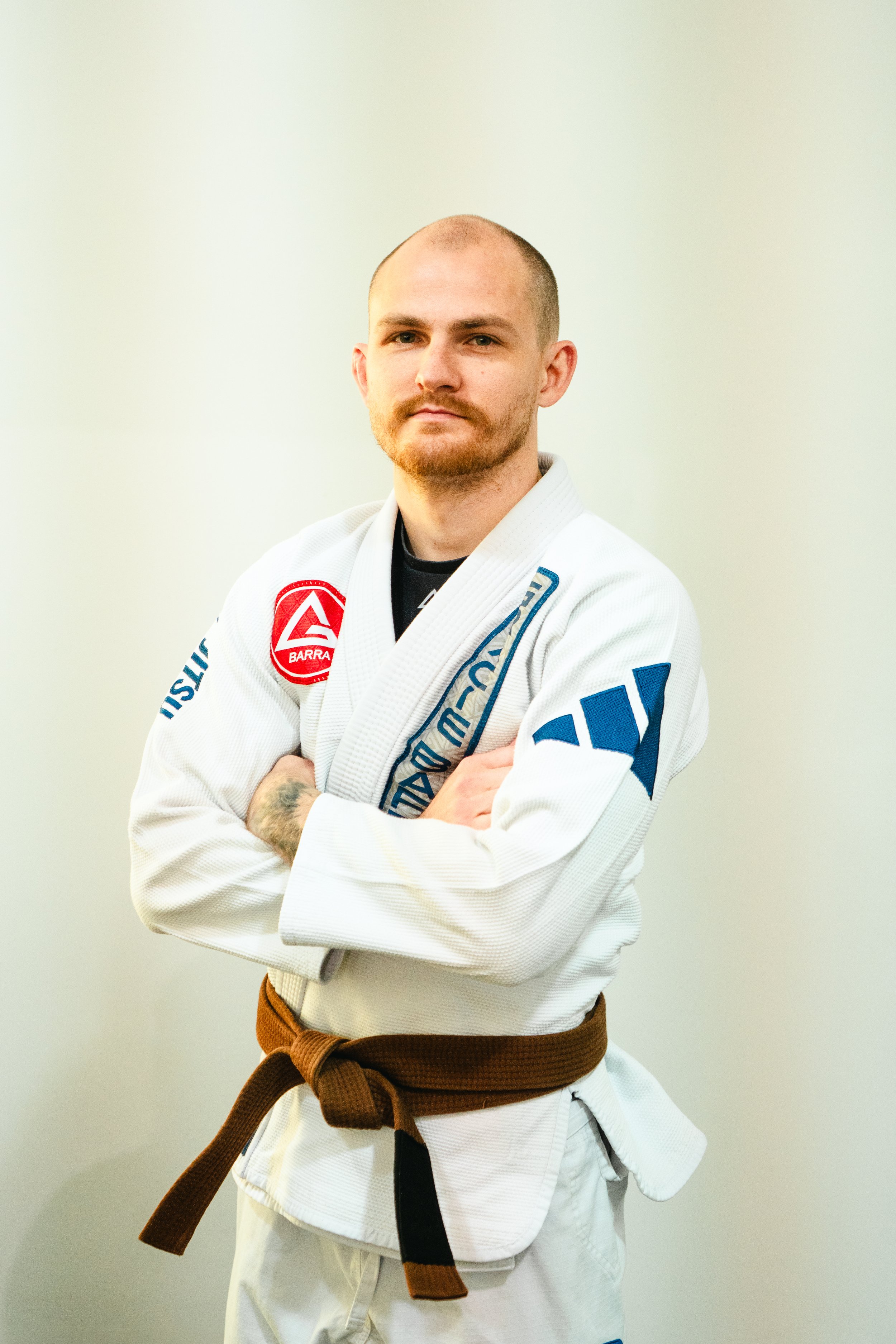Brazilian Jiu-Jitsu practitioner Coach Daniel at Gracie Barra Bryan wearing a white gi with patches and a brown belt, standing with arms crossed against a light background.