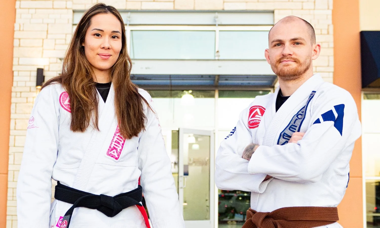 Coach Daniel and Professor Amanda of Gracie Barra Bryan wearing white Brazilian Jiu-Jitsu gis with colored belts, standing in front of the Gracie Barra Bryan gym.
