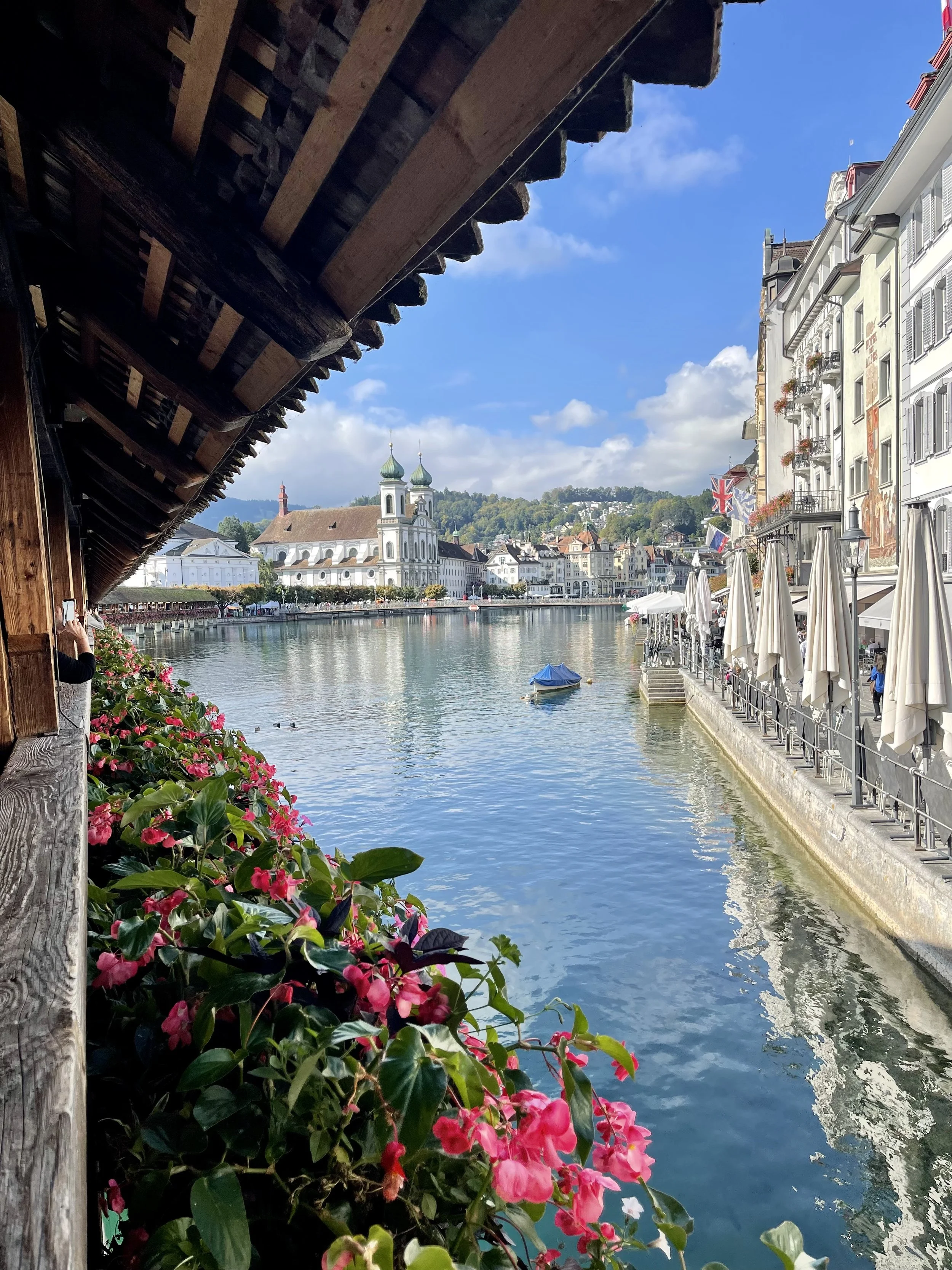 View of a river with a church and cityscape in the background, flowers and a balcony in the foreground, and a blue sky with some clouds.