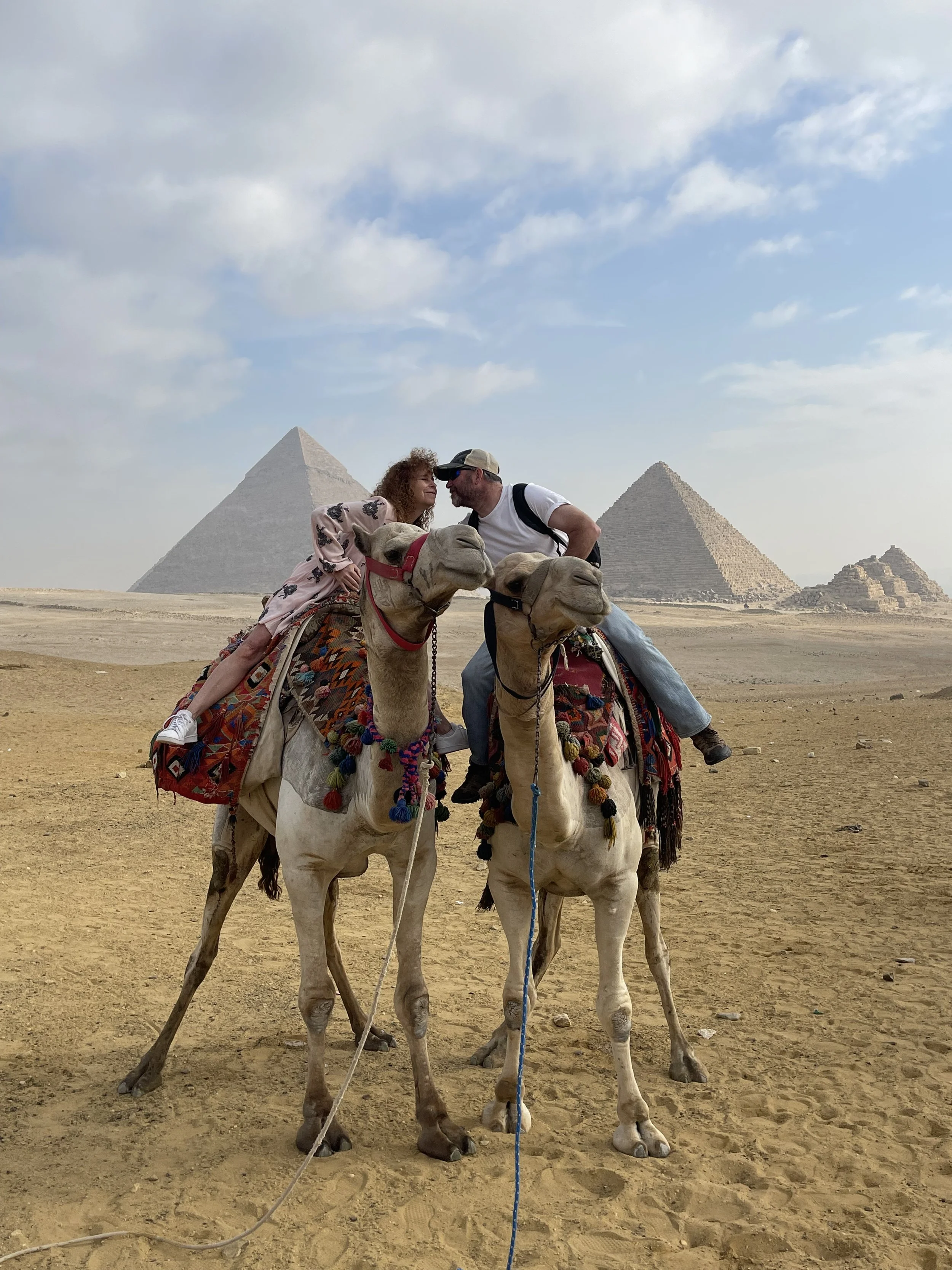 Two people riding camels in front of the pyramids in Egypt. The couple is leaning in towards each other, and the camels are decorated with colorful blankets and tassels.