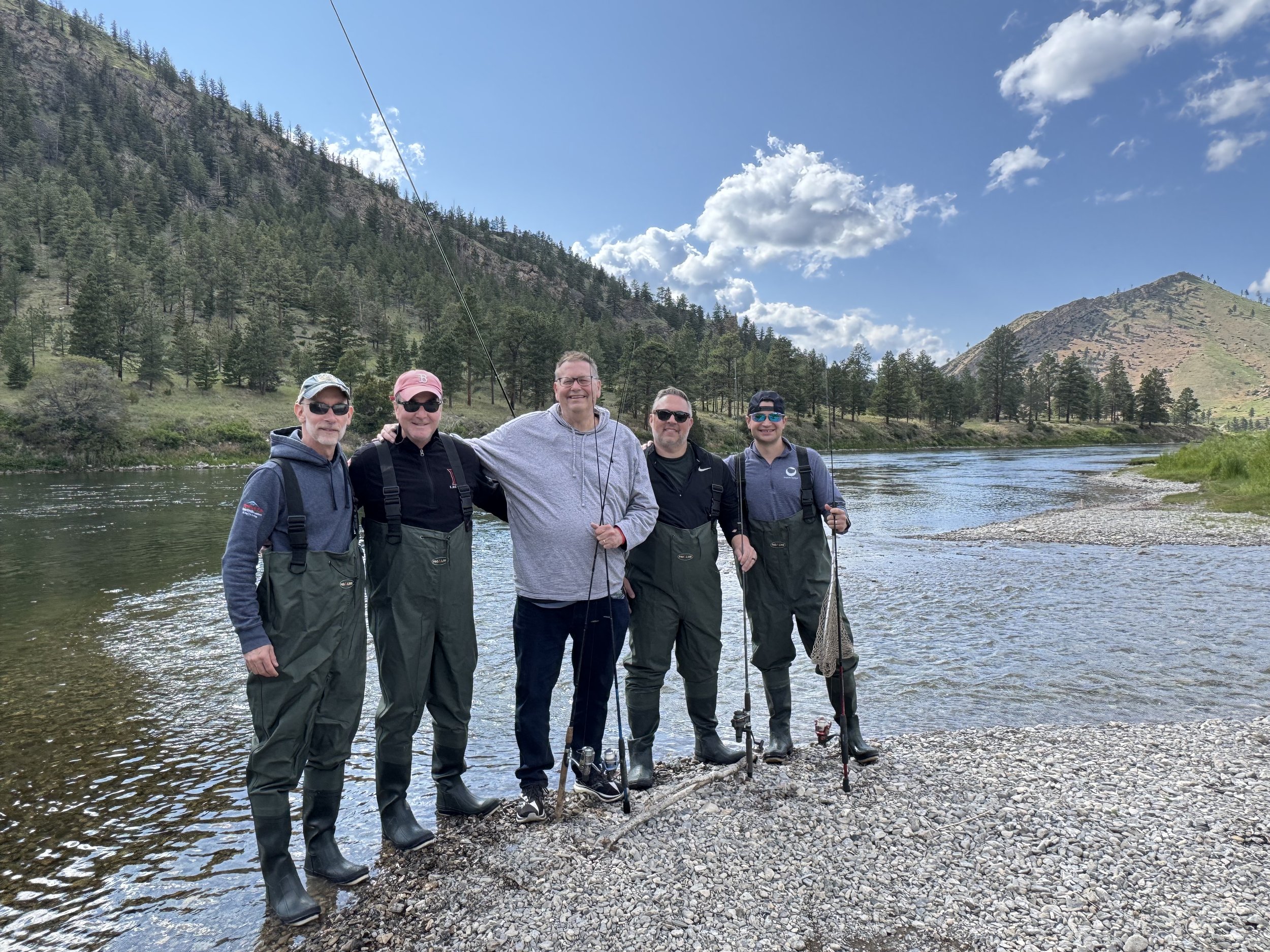 Group of five men standing on a rocky riverbank holding fishing rods, with a river, green forest, and mountains in the background on a partly cloudy day.