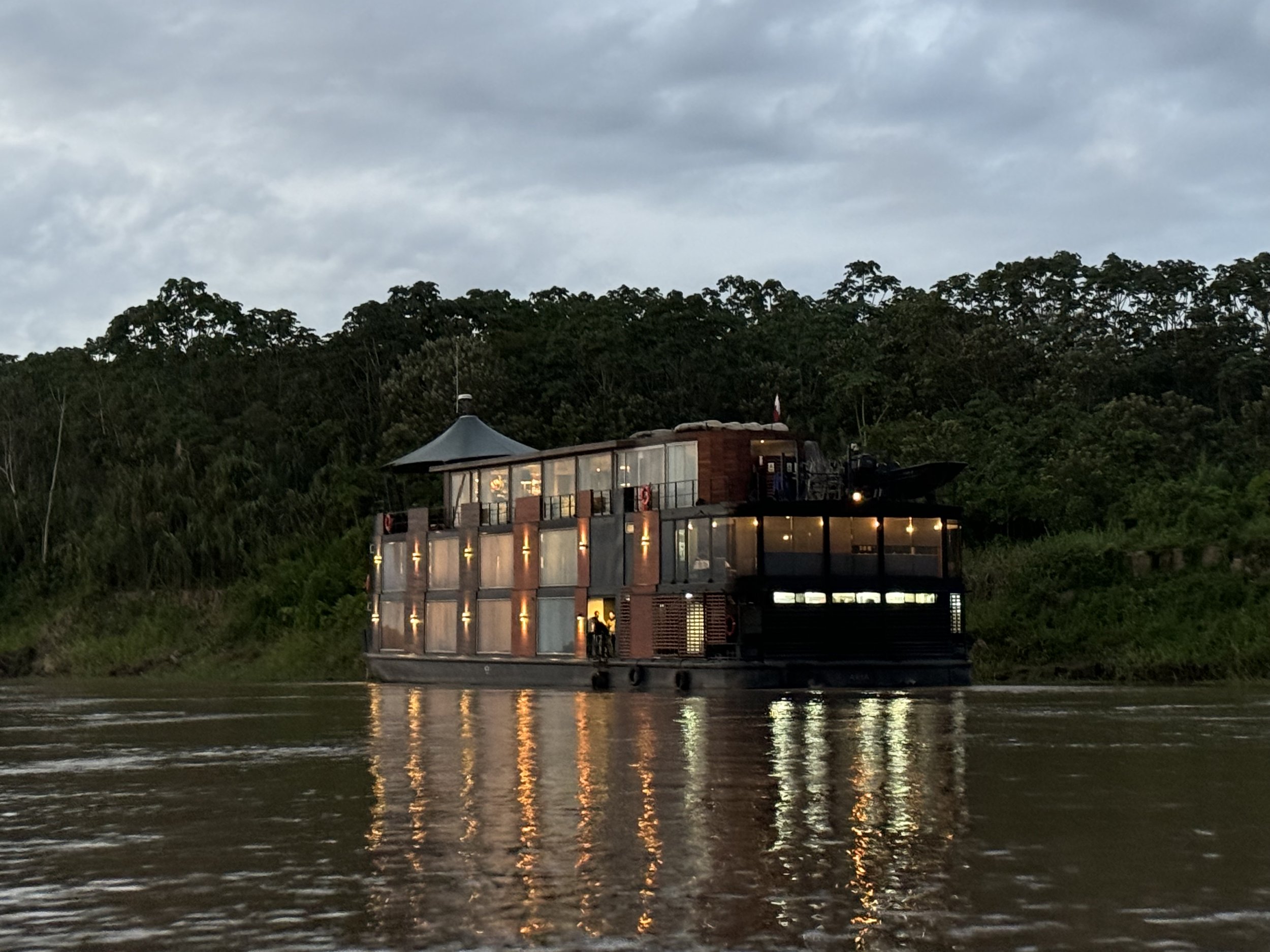 A houseboat with glass windows, a small upper deck, and an outdoor area with a canopy, floating on a river near a forested shoreline, during dusk or early evening.