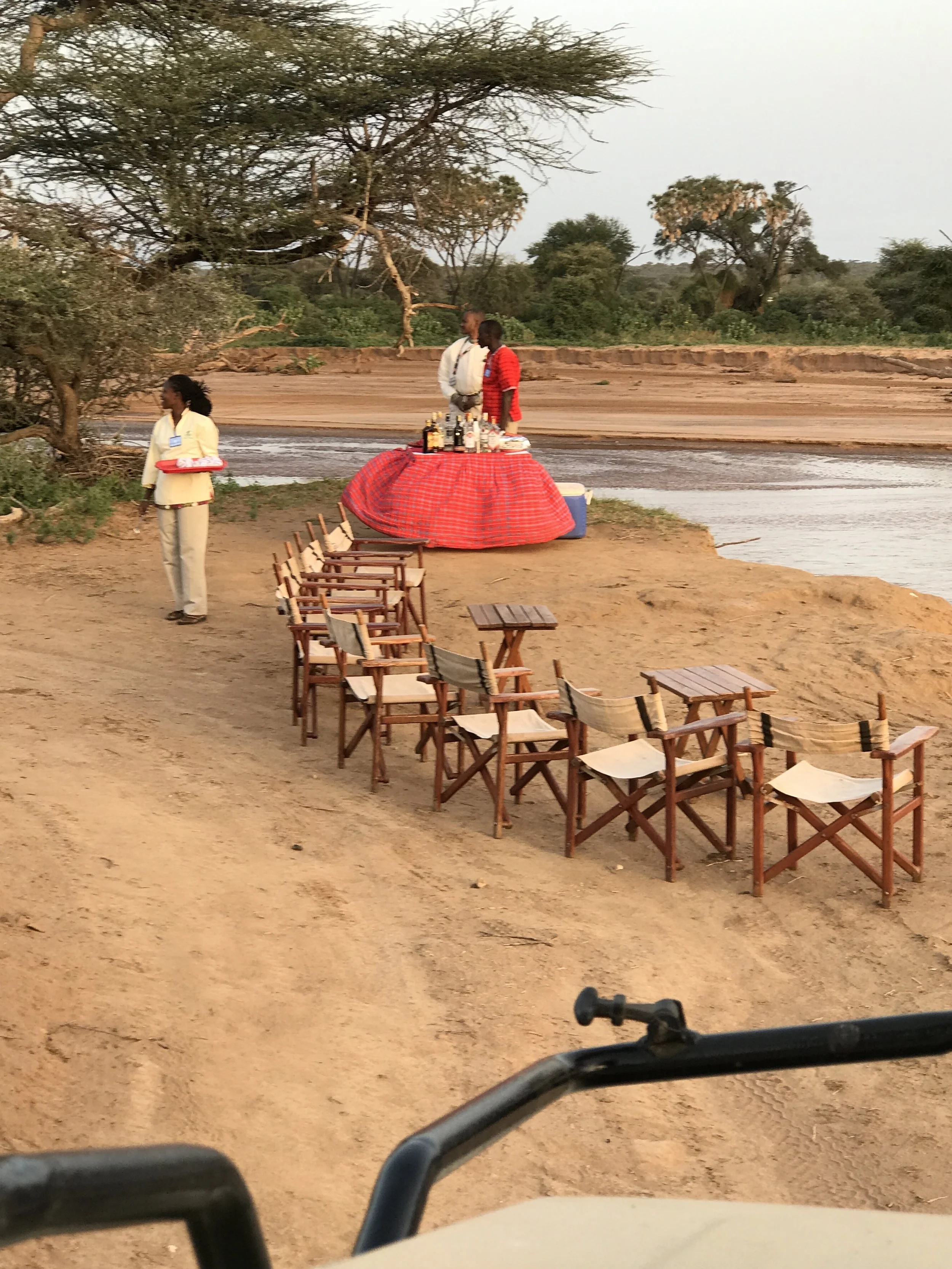 Outdoor scene with a river, trees, and a group of wooden chairs arranged on sandy ground, with a small bar setup covered by a red cloth, along with three people nearby, two of whom are standing by the bar.