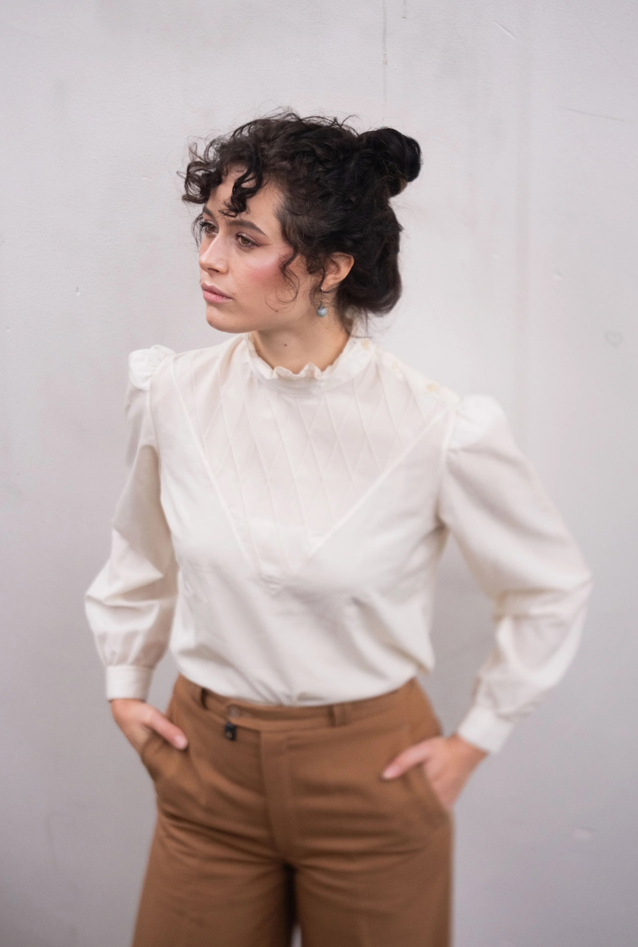 A woman with dark curly hair styled in a bun wearing a vintage white blouse with puffed shoulders and brown pants against a plain wall.