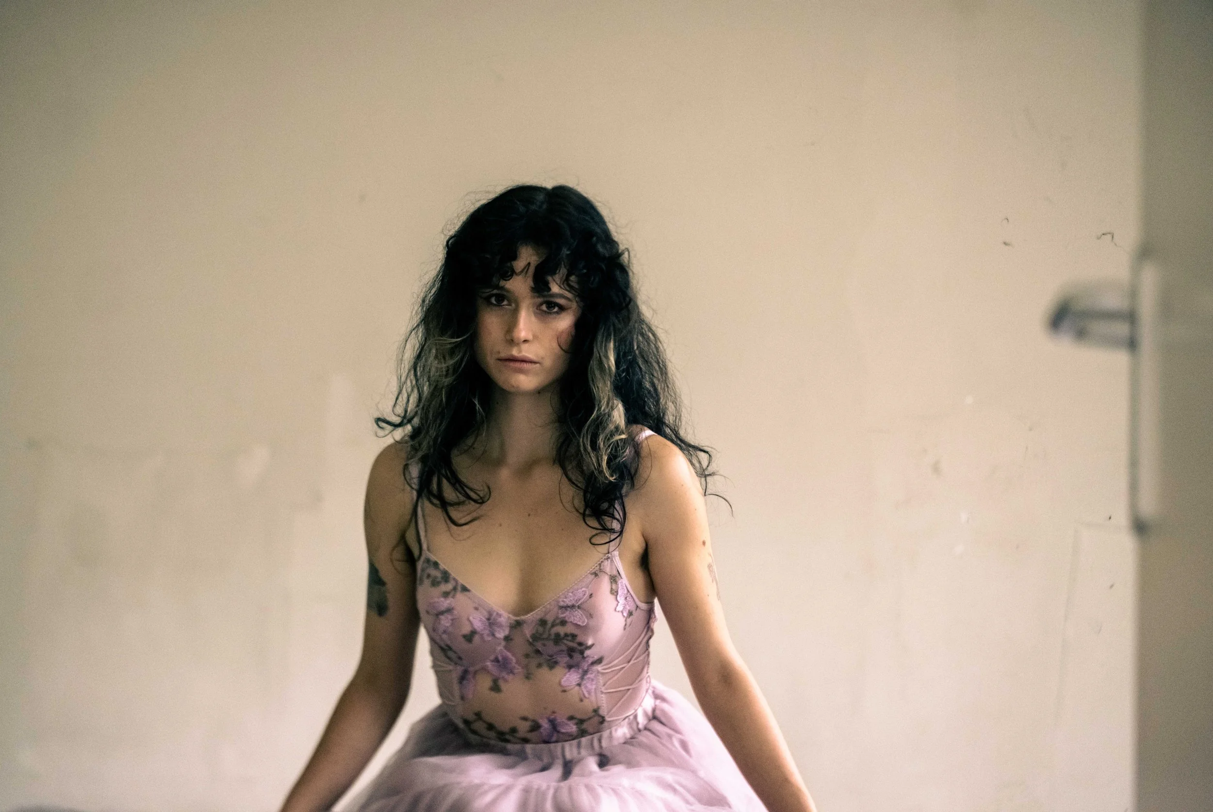 A woman with dark, curly hair and tattoos, wearing a pink floral dress, sitting against a plain wall with a serious expression.