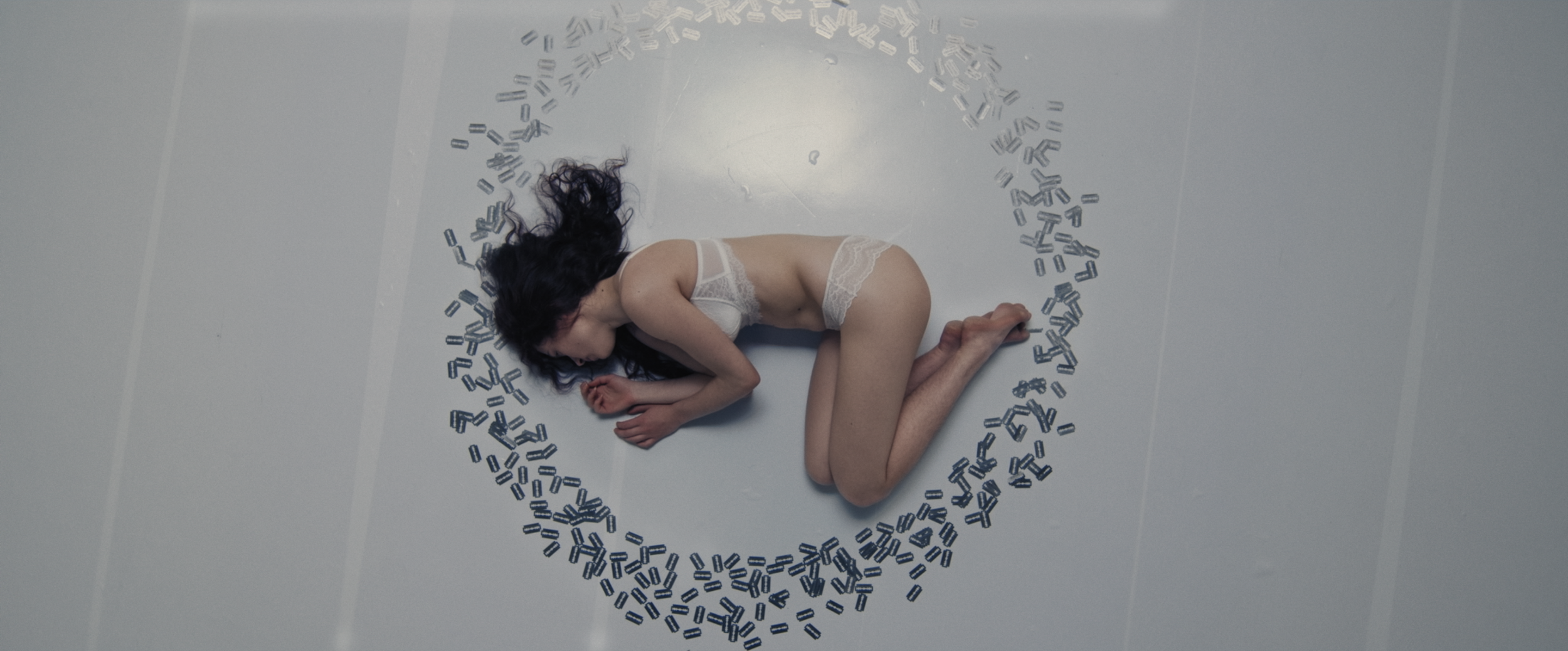 A woman with dark hair in white lingerie lying on her side on a plain gray floor, surrounded by a circle of scattered black and white pills.