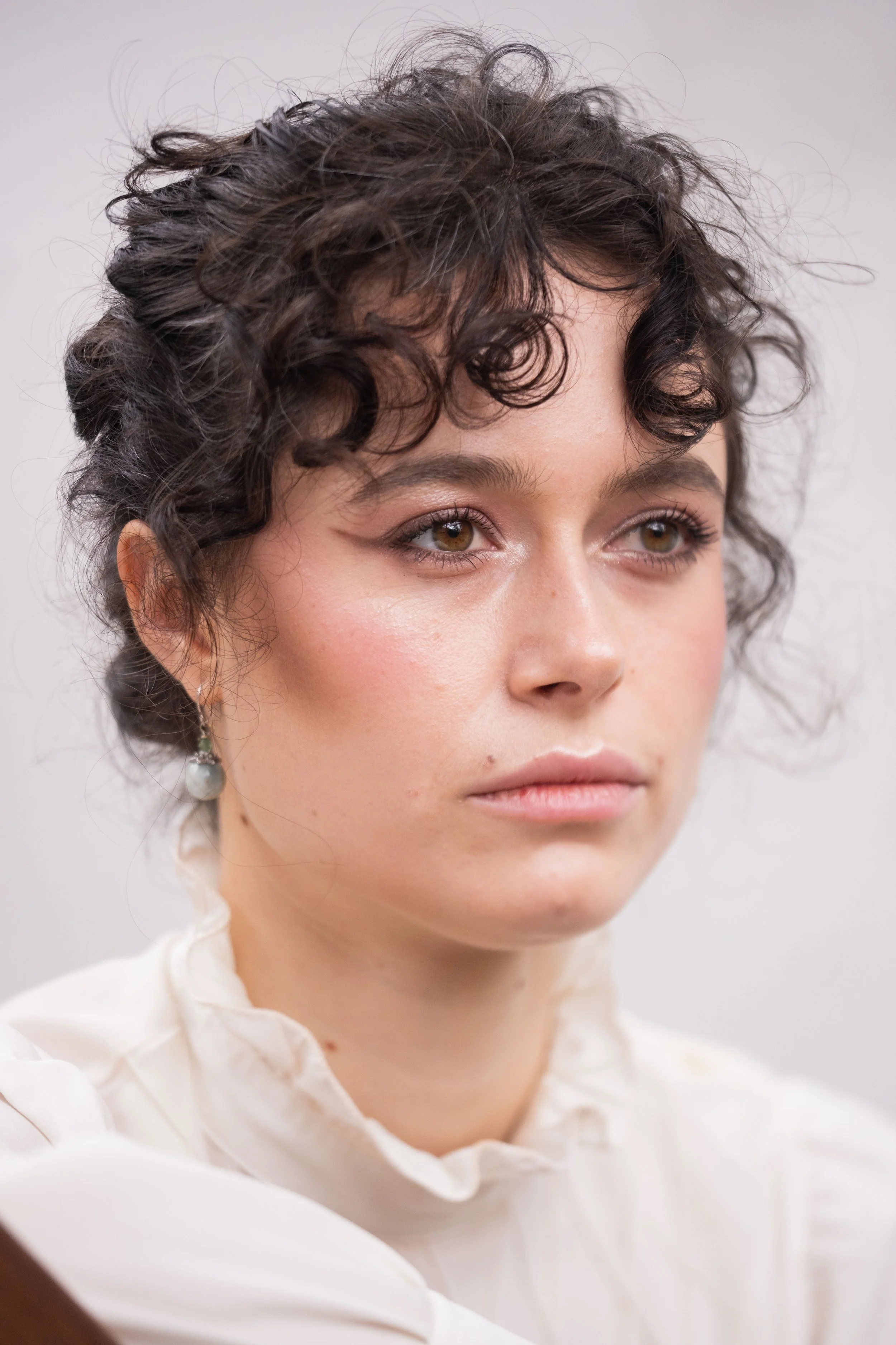 Close-up of a young woman with curly dark hair, wearing makeup, and a pearl earring, against a plain background.