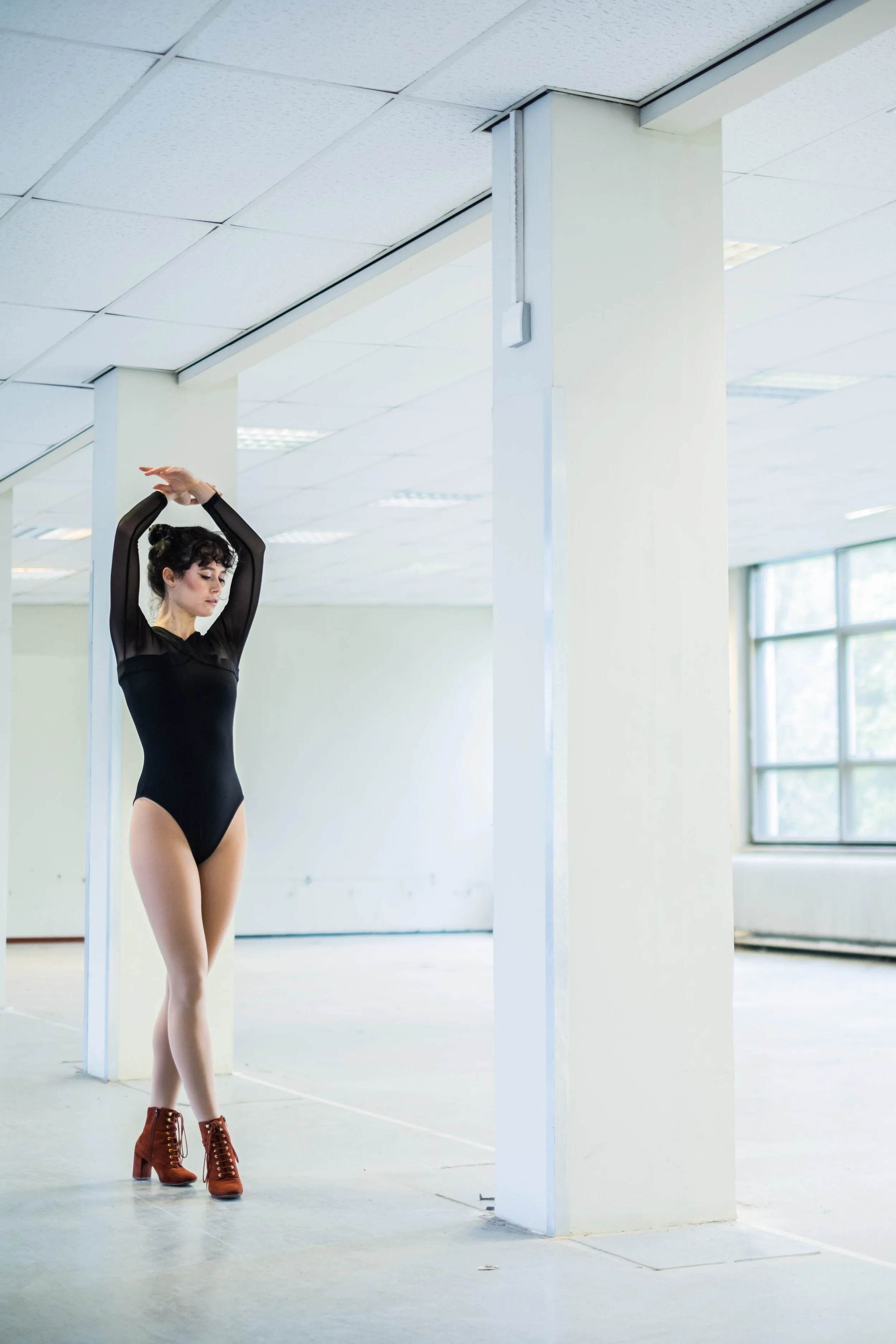 A woman in black dance attire and brown dance shoes standing in a spacious, white-walled dance studio with large windows, stretching her arms above her head.