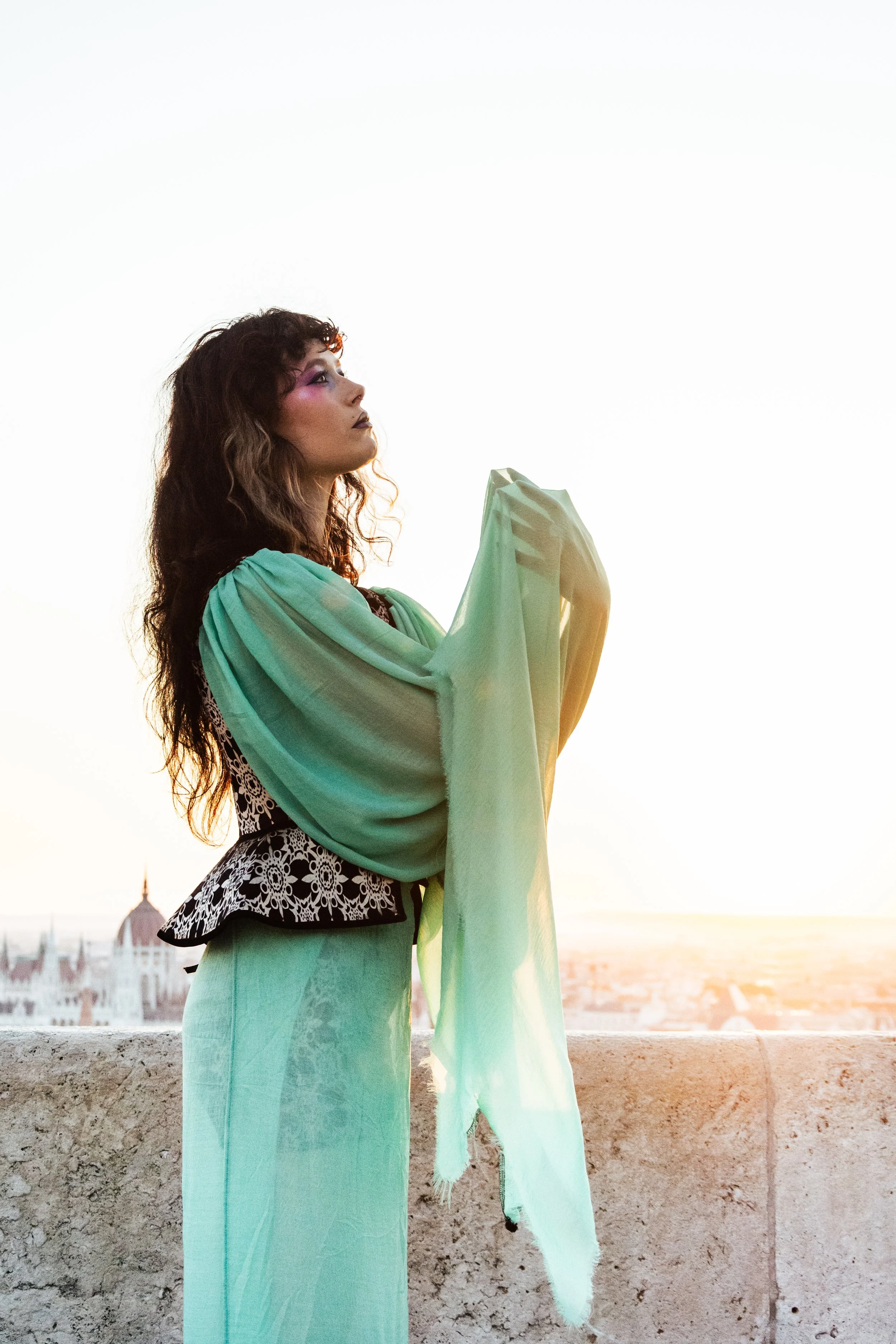 A woman standing on a rooftop at sunset, wearing a green shawl and a patterned black-and-white top with matching green pants.