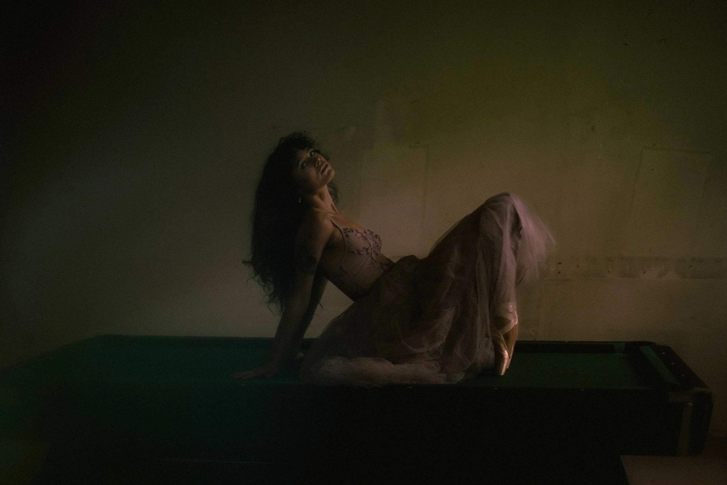 A woman with long dark hair sitting on a pool table in a dimly lit room, wearing pointe shoes, looking upward.