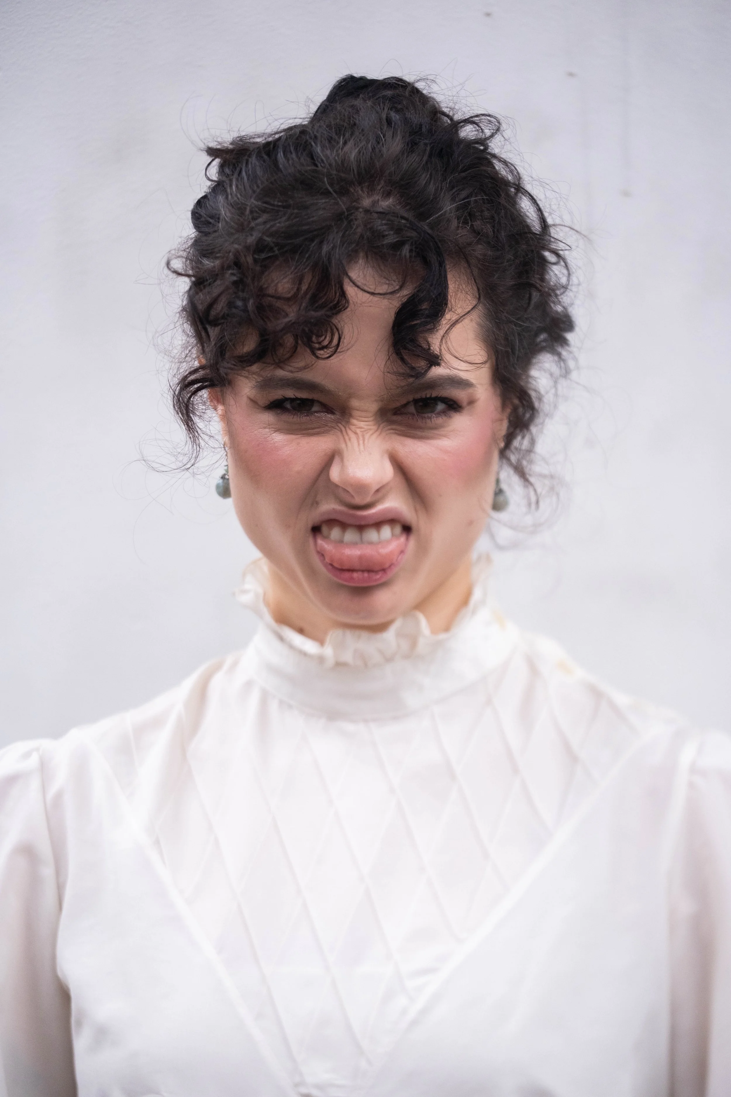 A woman making a face with her tongue out and her eyes squinted, wearing a white high-neck blouse.