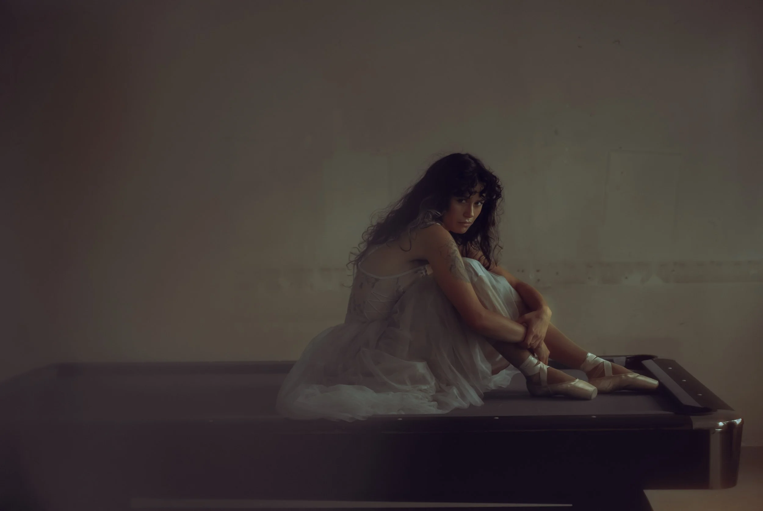 A woman in a ballet tutu sitting on a massage table in a dimly lit room, looking towards the camera with her arms wrapped around her knees.