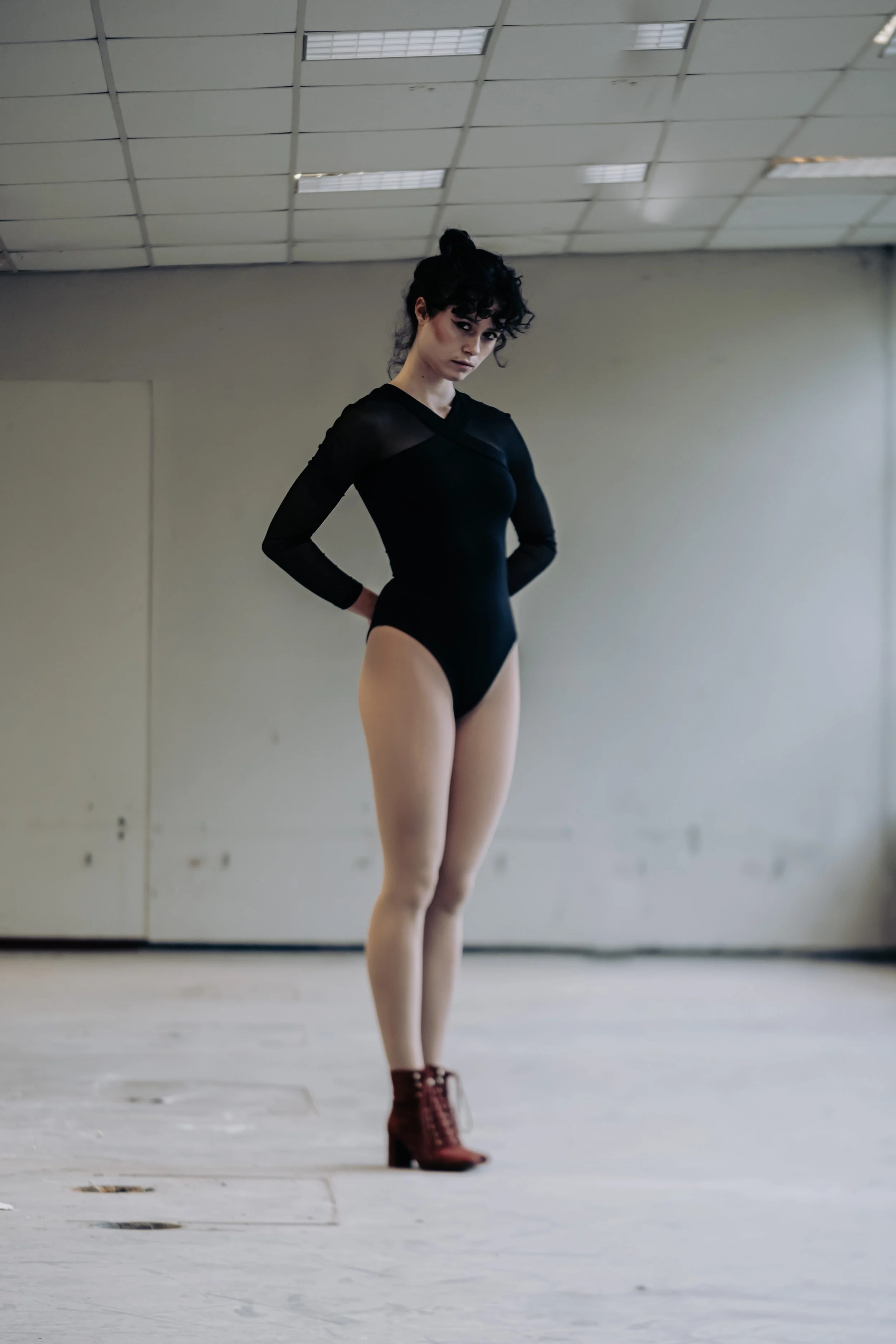A young woman with curly black hair styled in a bun, wearing a black long-sleeve leotard and burgundy ankle boots, standing in an empty indoor space with a drop ceiling and plain walls.