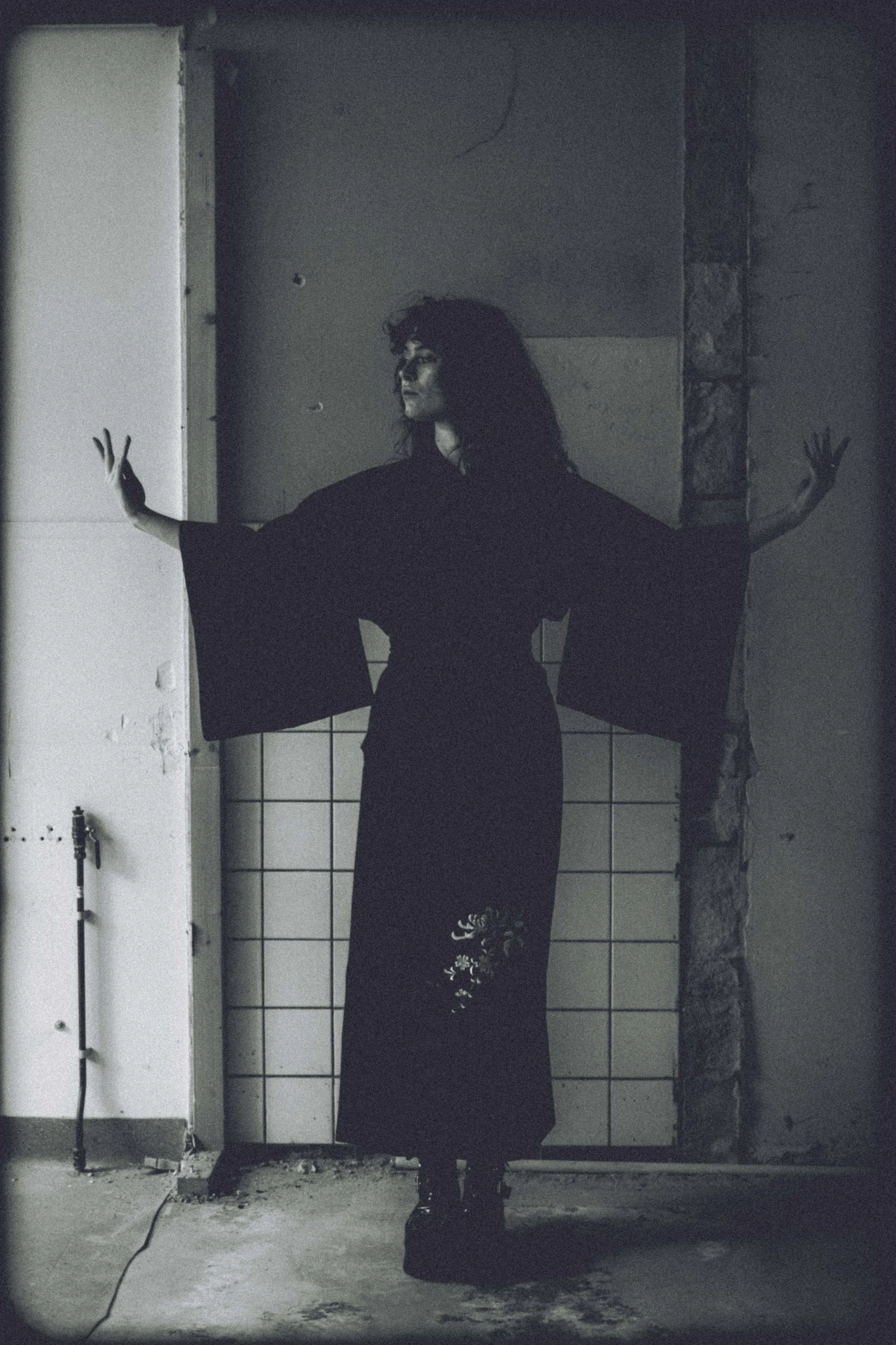A woman with curly hair in a black kimono or dress with floral embroidery, standing with arms outstretched in a dimly lit, abandoned room with worn walls and tile background.
