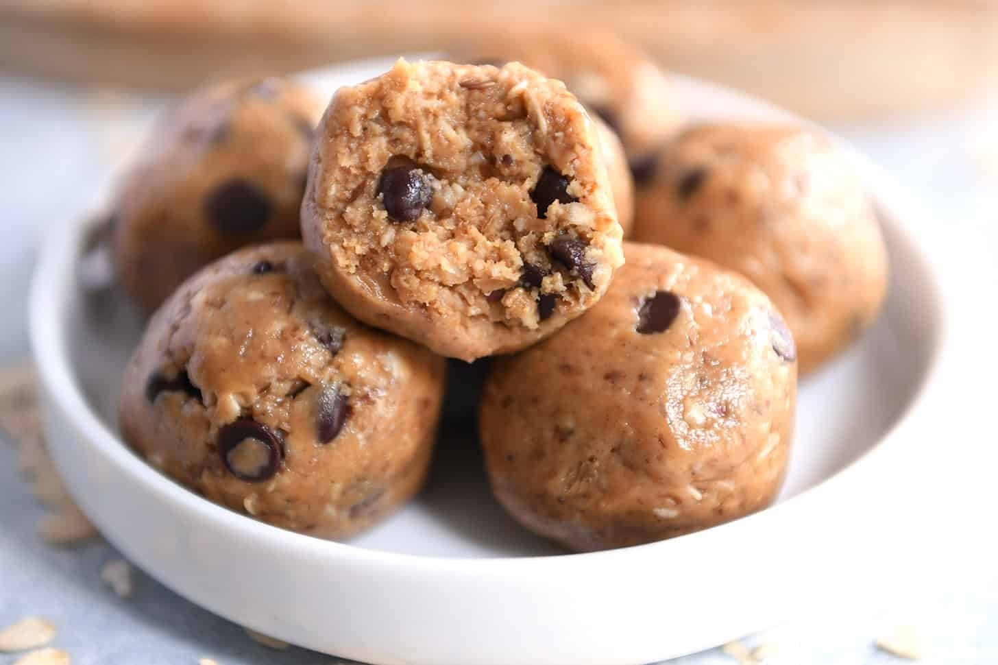 Close-up of chocolate chip cookie dough balls in a white bowl, with one bitten to reveal texture and chocolate chips inside.