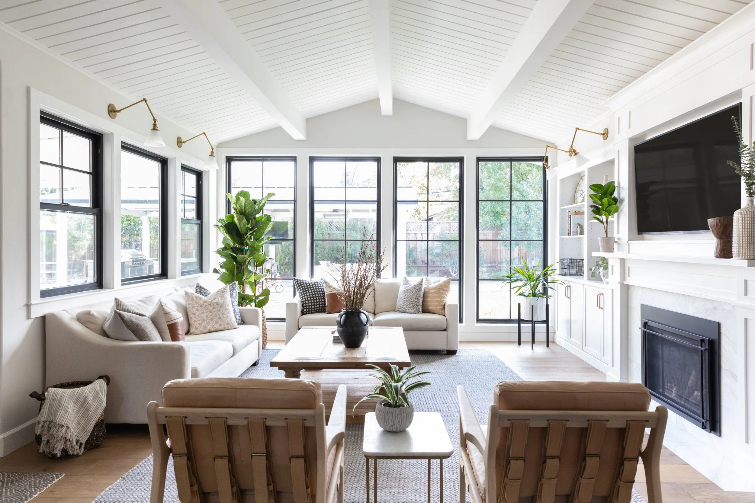 Bright living room with white sofas, wooden coffee table, and large windows with black frames, decorated with potted plants and a built-in white cove with a TV and fireplace.