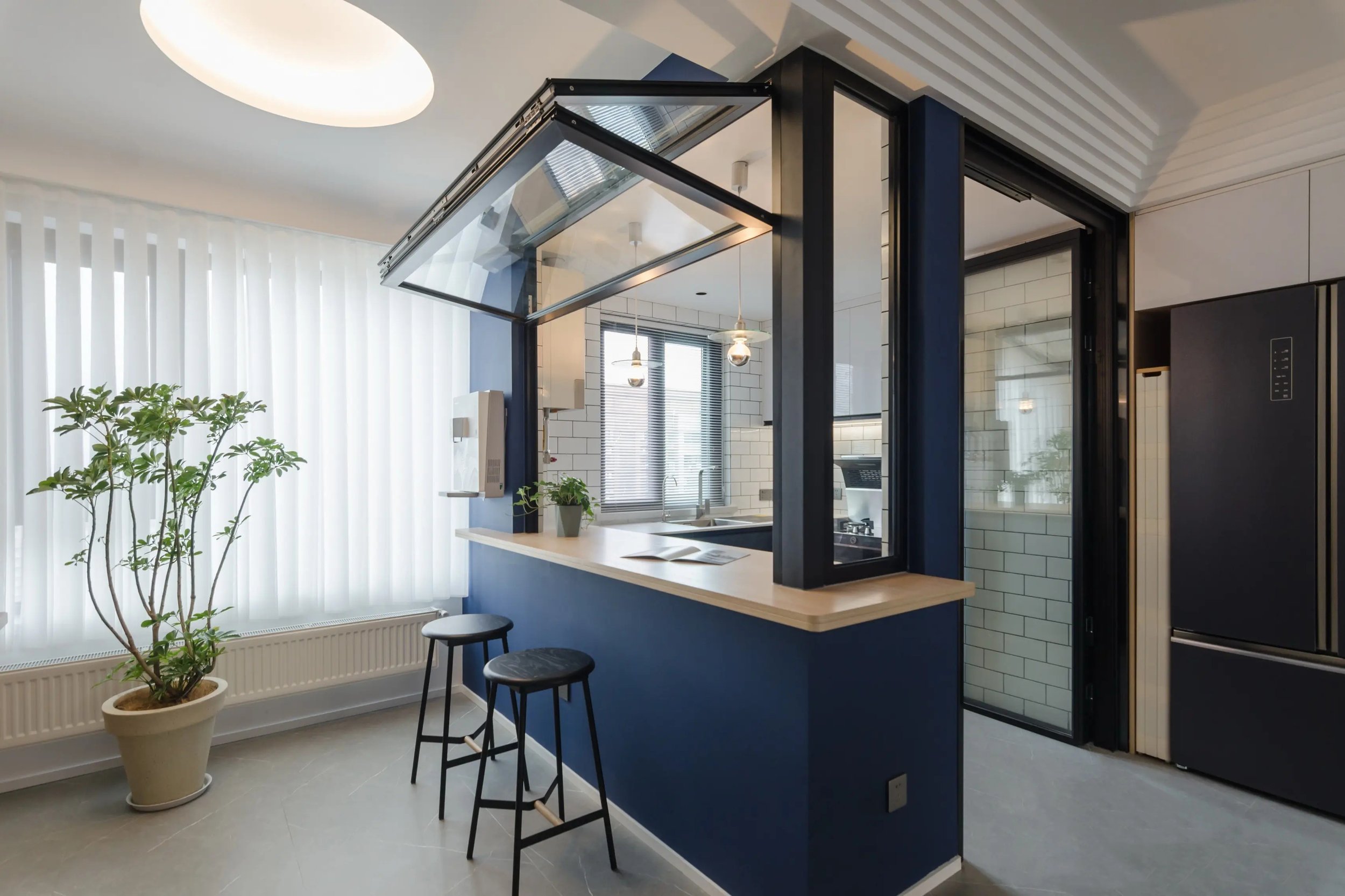 Modern kitchen with a peninsula and two bar stools, large potted plant near a window with white blinds, black and white color scheme, glass door leading to a pantry or utility area, and pendant lights over the sink.