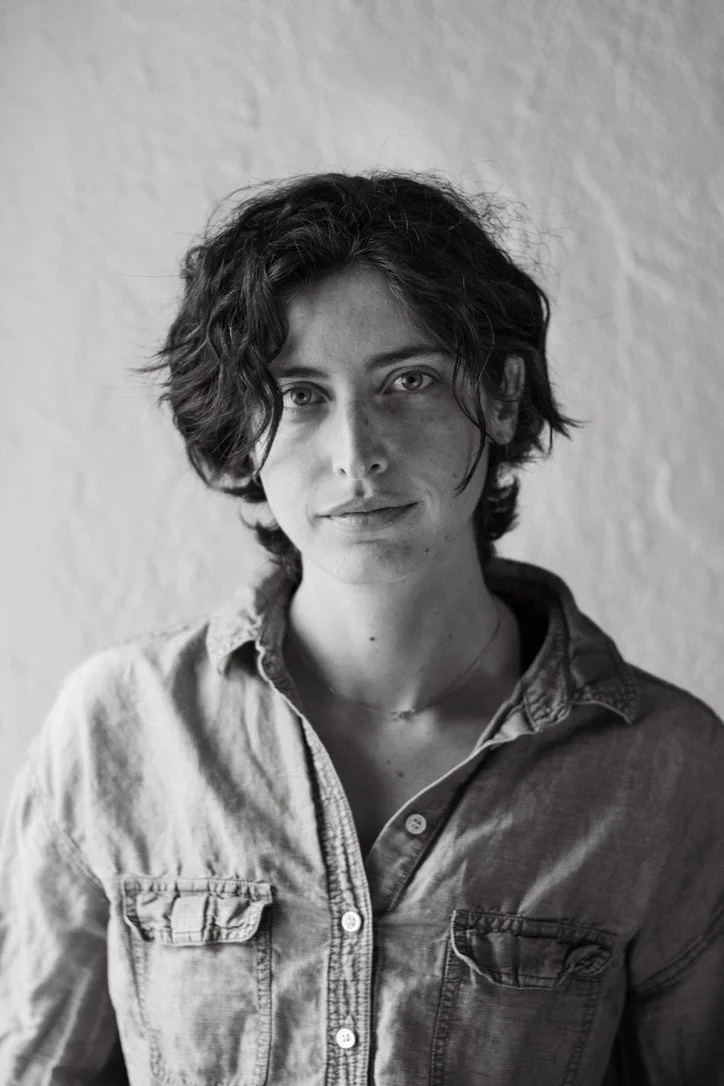 Black and white portrait of a young woman with short, wavy hair, wearing a button-up shirt, standing against a plain textured background.