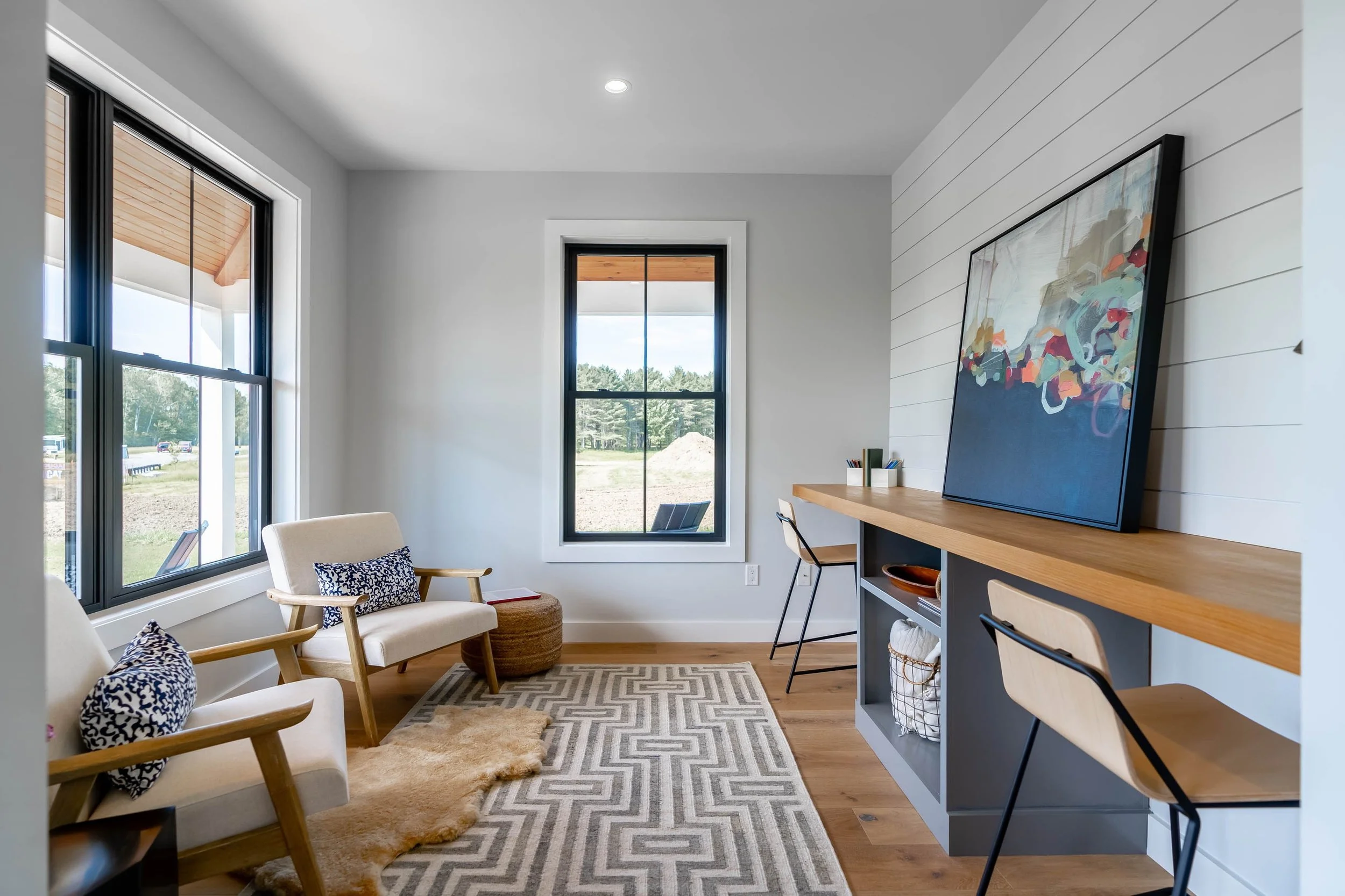 Modern living room with large windows, white walls, patterned area rug, armchairs with blue patterned pillows, and a wooden writing desk with a large colorful abstract painting on the wall.