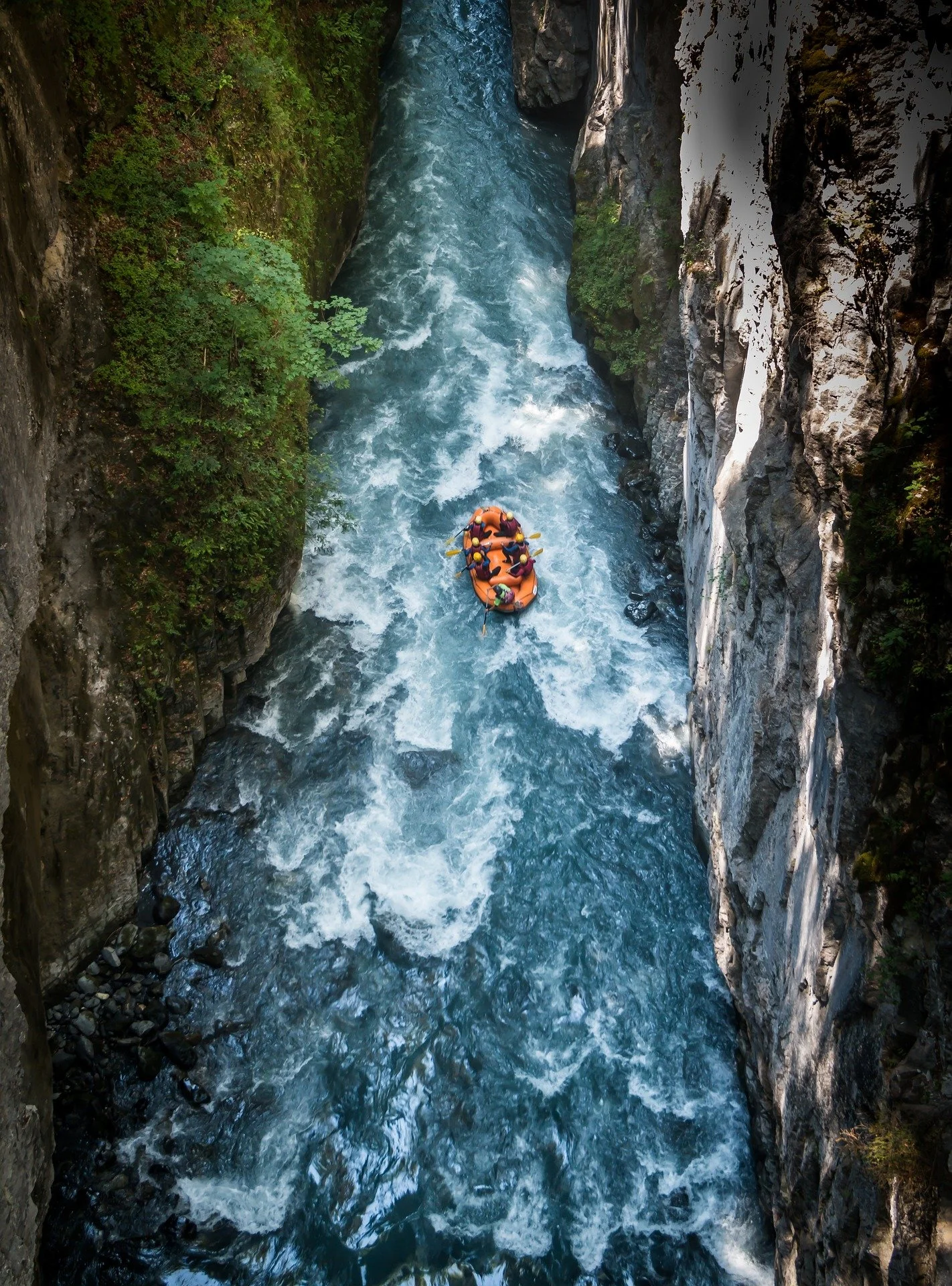 A group of people white-water rafting in a narrow canyon with steep rocky walls and some greenery.