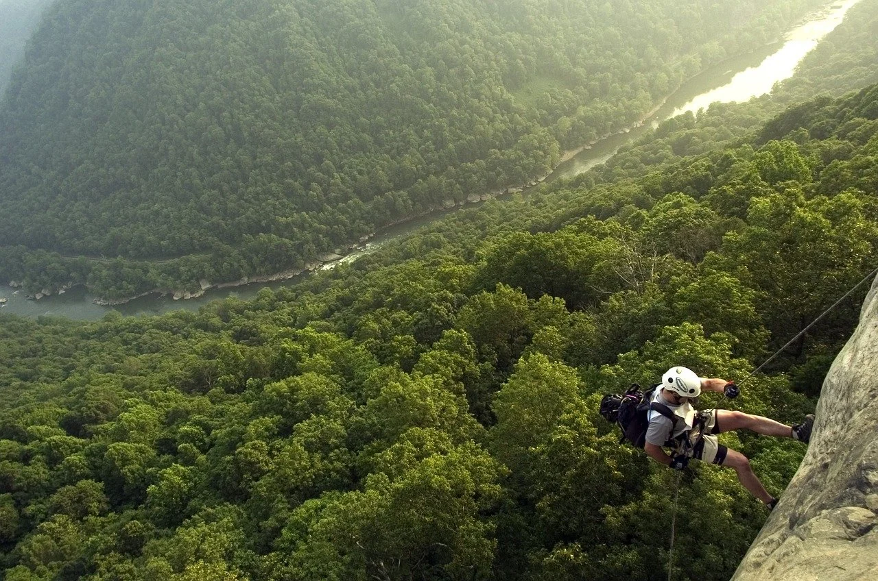A person rock climbing on a steep cliff in a lush green forest with a river flowing through the valley below.