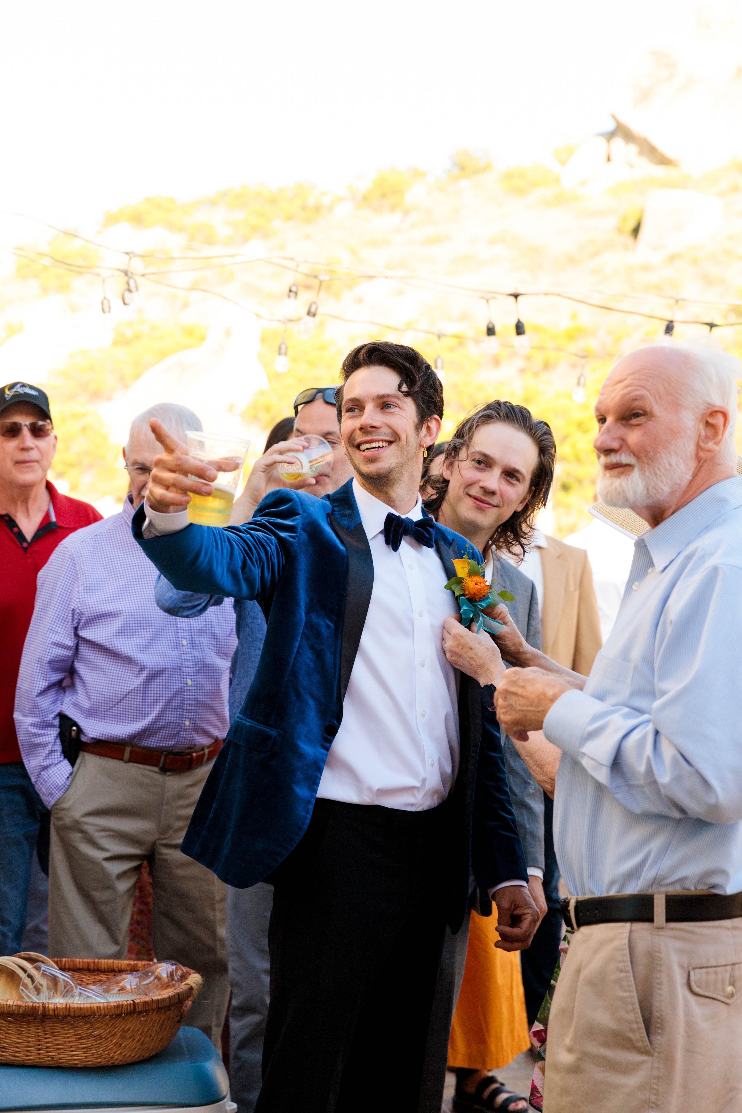 A group of people gathered outdoors, celebrating, with a man in a blue velvet tuxedo taking a selfie, surrounded by friends in formal attire. The background shows string lights and a sunny, outdoor setting.