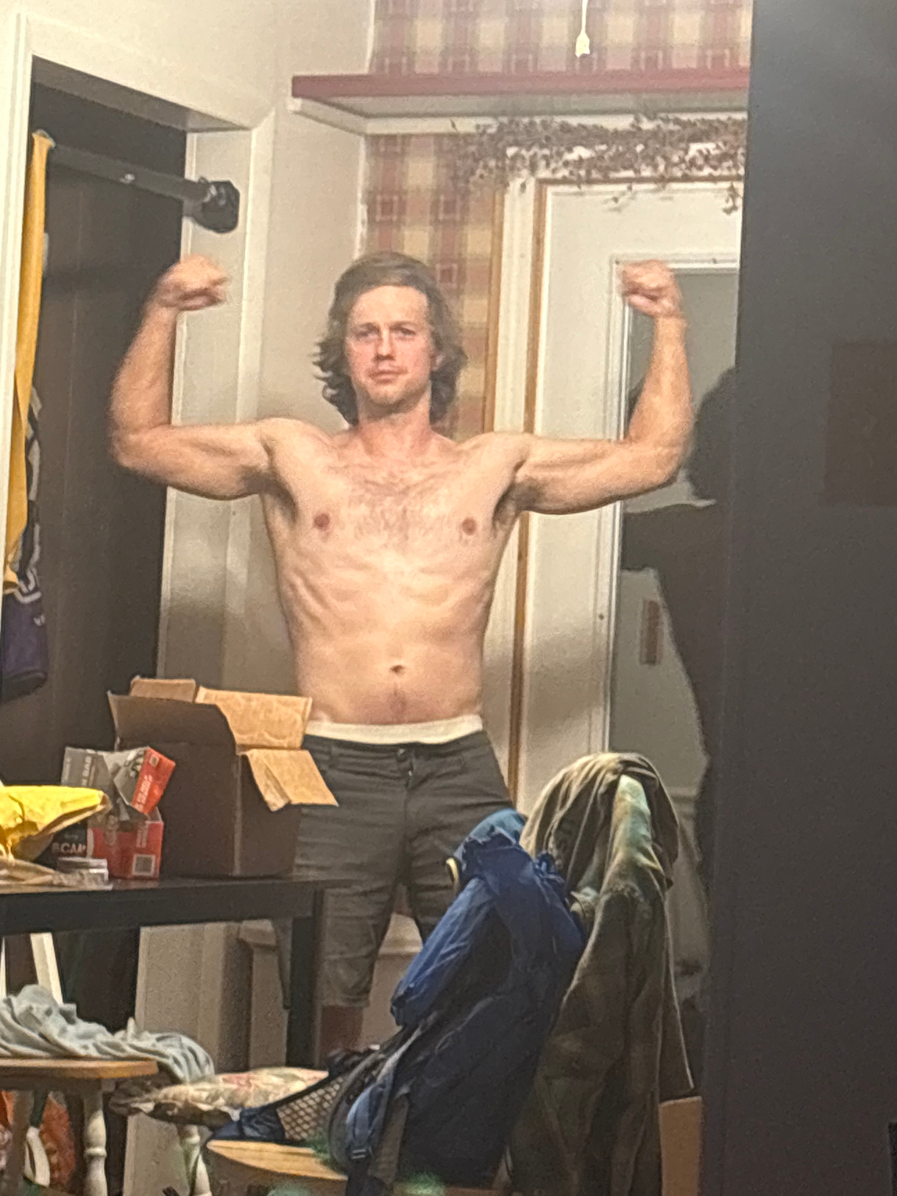 A shirtless man with long hair flexes his biceps in front of a mirror. The room contains various items and furniture, including a table with snacks and a chair with a backpack on it.
