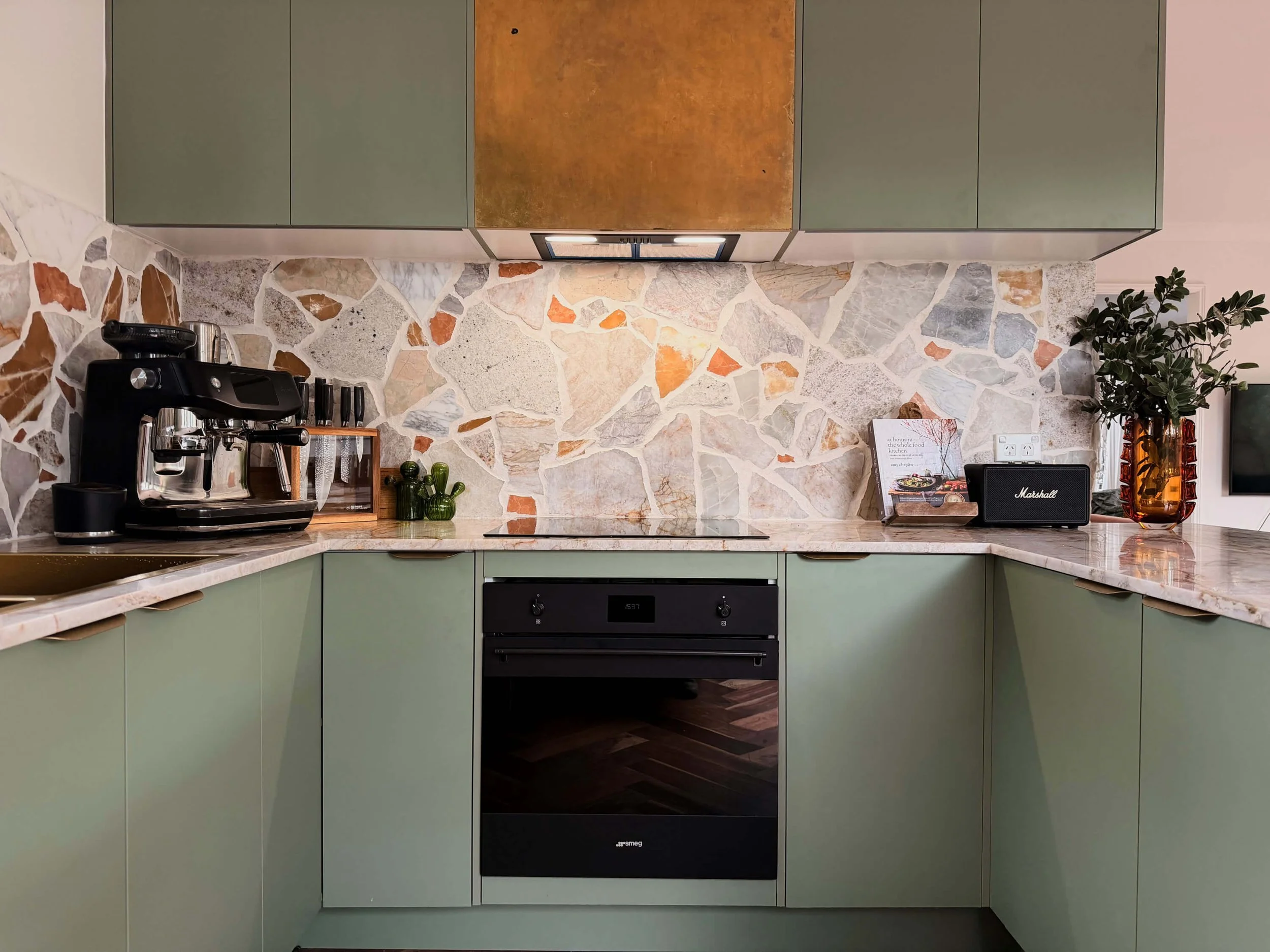 A kitchen with green cabinets, a stone tile backsplash, and a black oven. There is a coffee machine, green vases, and a cookbook on the counter.