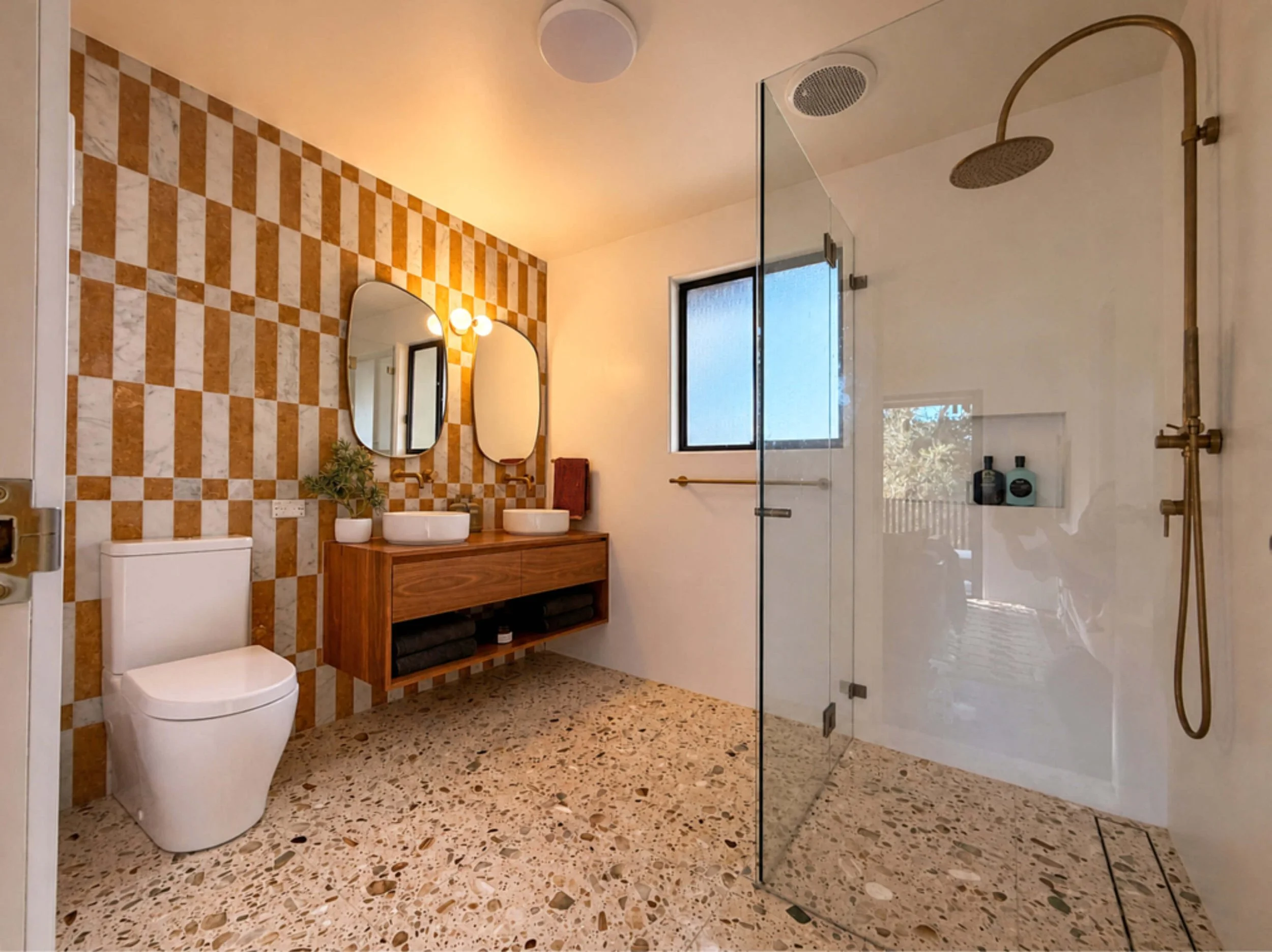 Modern bathroom with terrazzo floor, checkered wall tiles, two oval mirrors above a wooden vanity with two vessel sinks, a wall-mounted toilet, a frosted glass window, and a walk-in shower with a brass rain showerhead and hand shower.