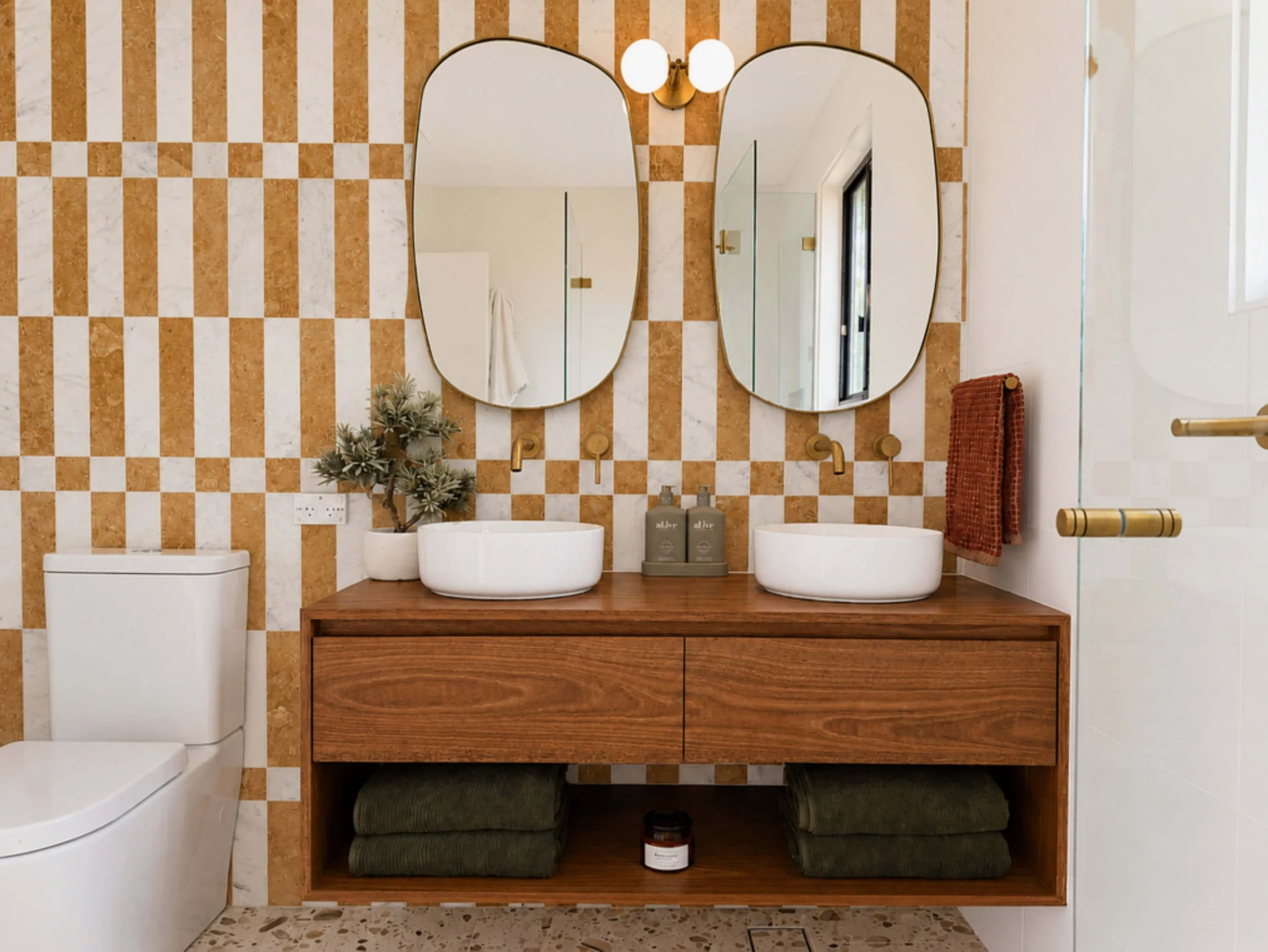 A modern bathroom with a double sink vanity, oval mirrors, a patterned wallpaper, and towels hanging on the side.
