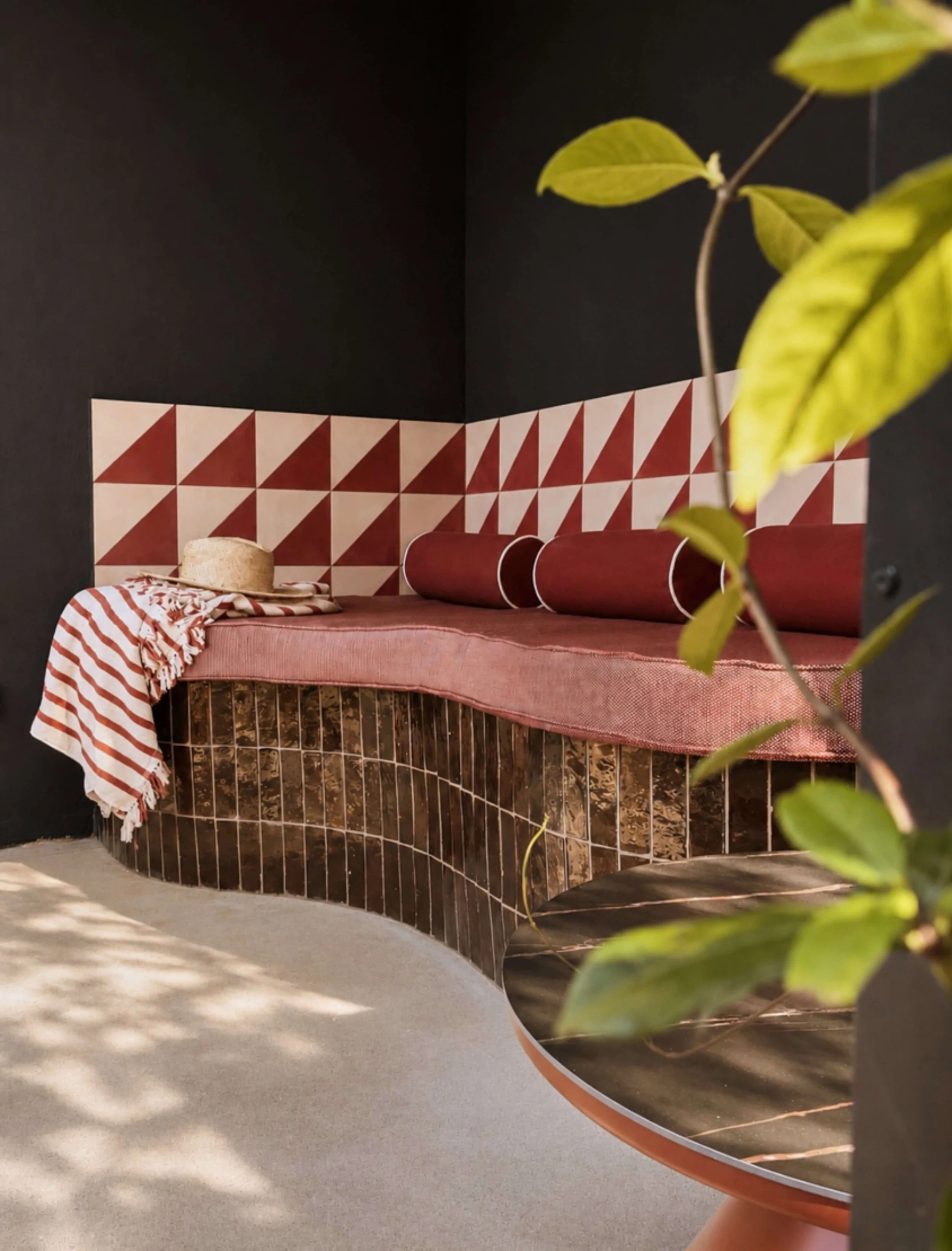 Indoor corner with black walls, red and white tiled wall, curved brown tiled counter with a pink tablecloth, a striped cloth, straw hat, and a green leafy plant in a round framed mirror.