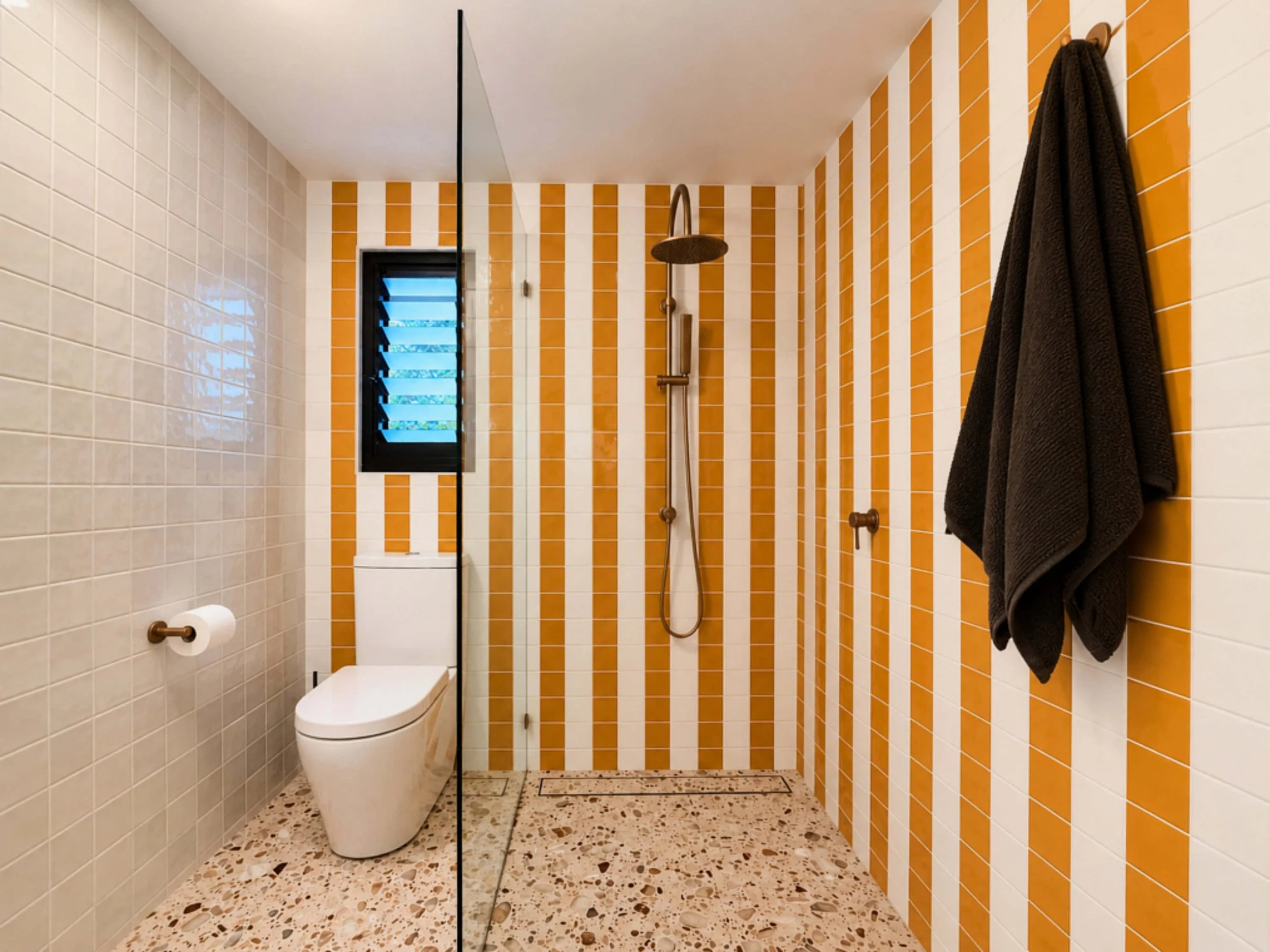Modern bathroom with orange and beige striped tile walls, a black-framed window with louvered glass, a ceiling-mounted showerhead, a grab bar, a toilet, a toilet paper holder, and a dark towel hanging on the wall.
