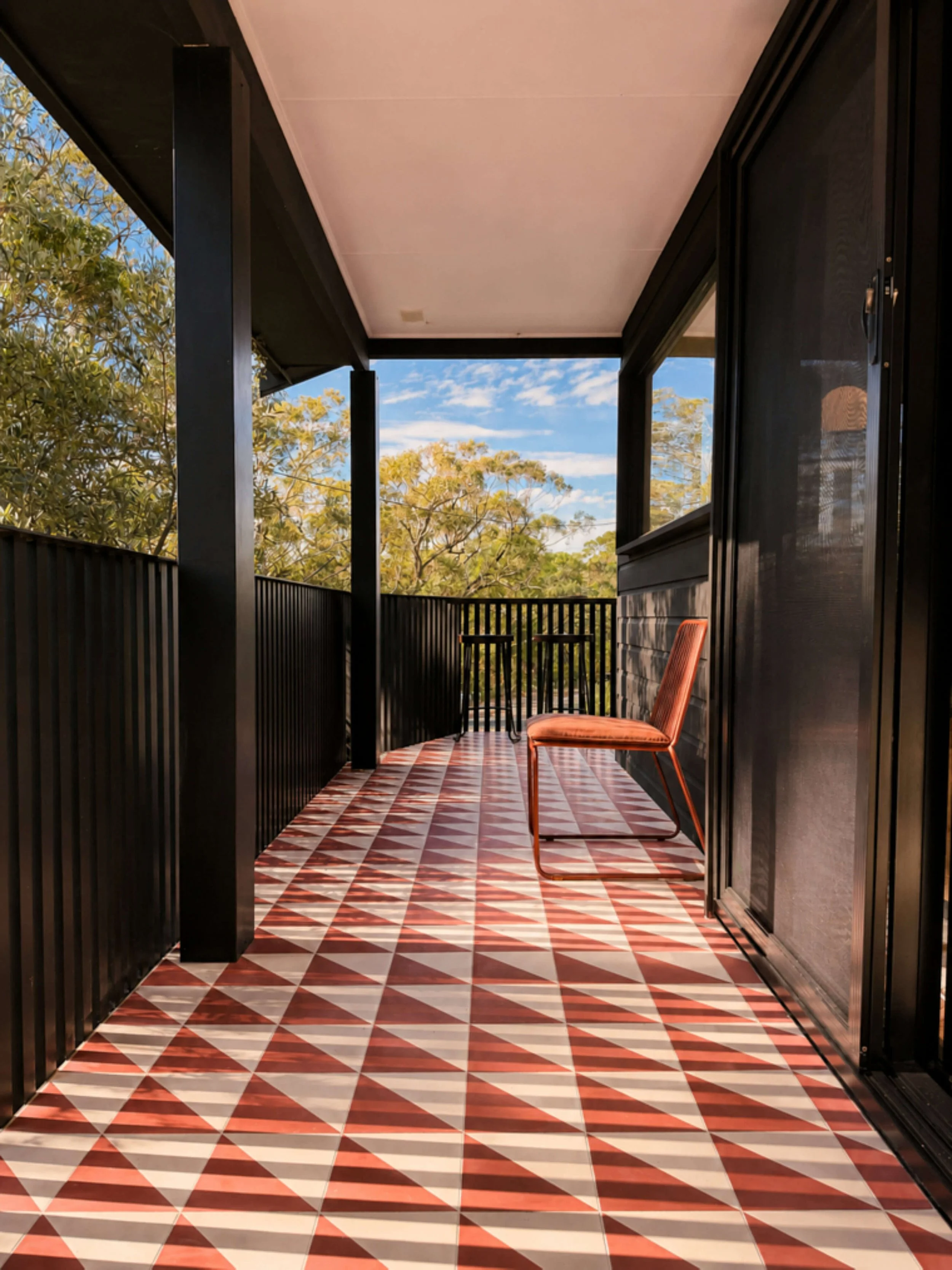 View of a covered balcony with a red and white diamond patterned tile floor, black railing, black support beams, a black wall with a sliding door, and a view of blue sky with scattered clouds and trees in the background.