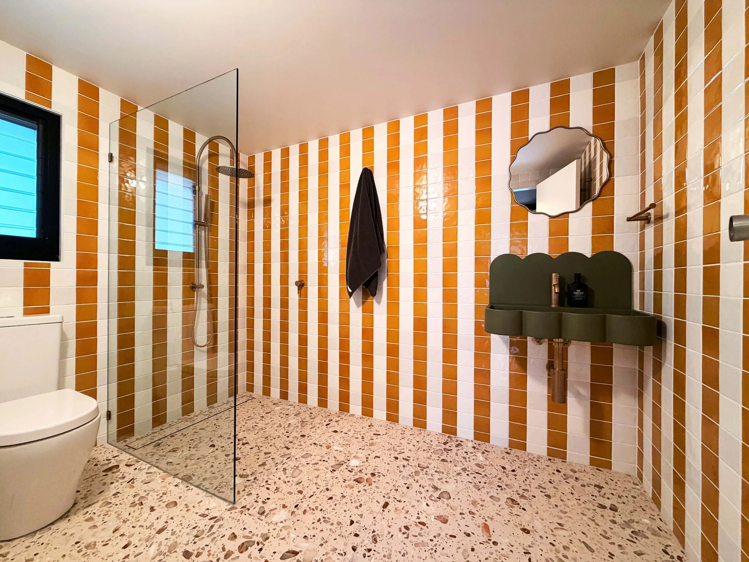 Modern bathroom featuring orange and white striped ceramic wall tiles, terrazzo flooring, a glass shower enclosure, a wall-mounted mirror, a gray sink with a soap dispenser, and a black towel hanging on the wall.