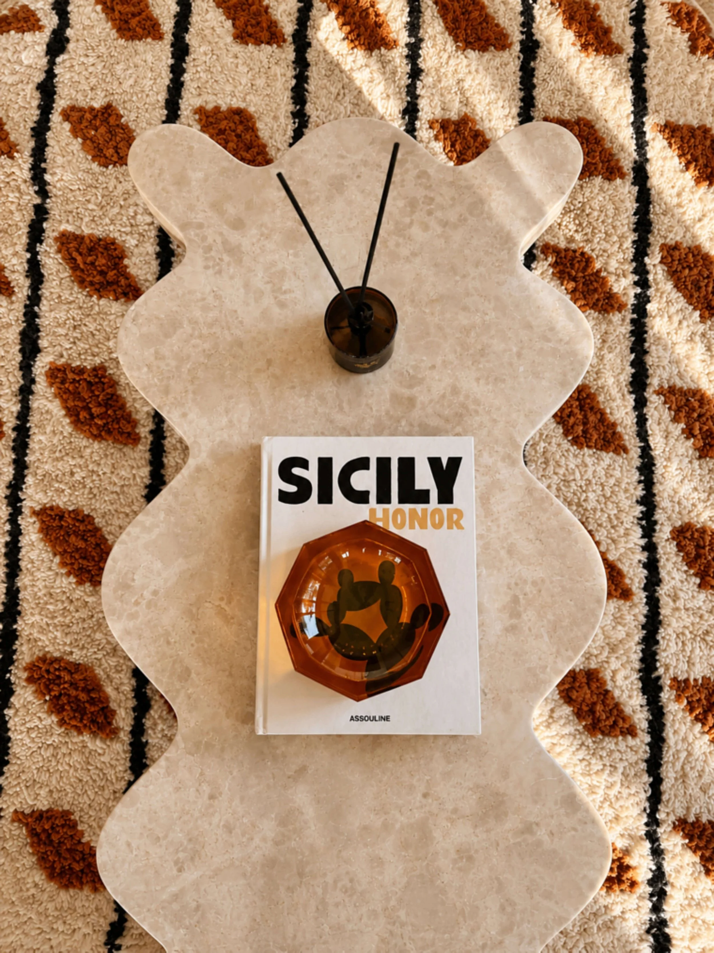 Close-up of a floral patterned carpet with a marble table on top, holding a black candle with black wicks, a book titled "SICILY HONOR," and a small amber glass dish with a silhouette of a dog or wolf inside.