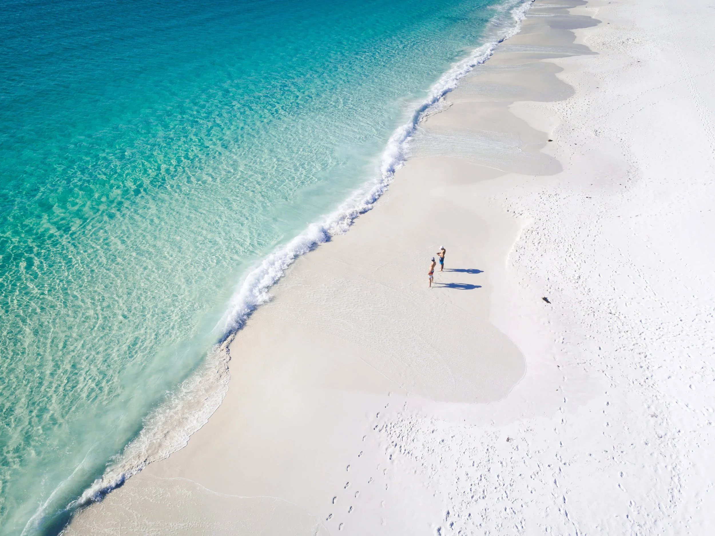 Aerial view of a sandy beach with clear turquoise waters, three people standing near the shoreline, facing the ocean.