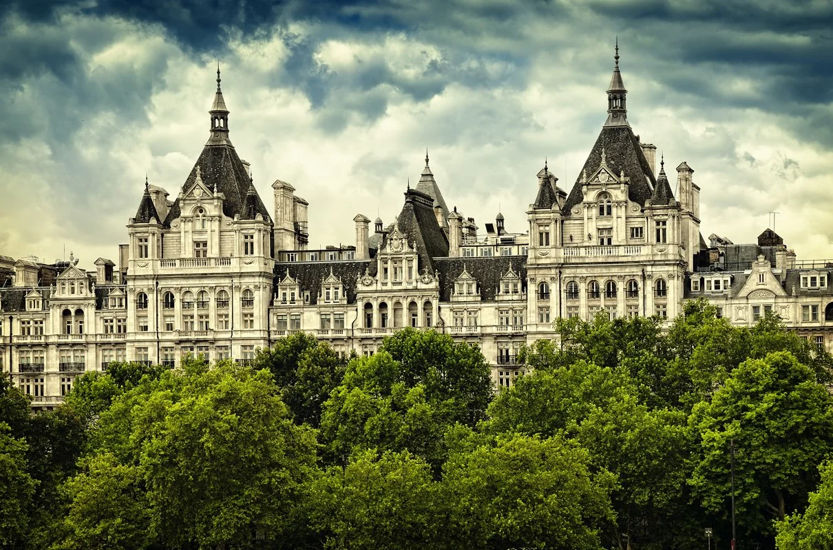 A large, ornate castle-like building with multiple turrets, spires, and decorative architecture, partially obscured by lush green trees in the foreground, under a cloudy sky.