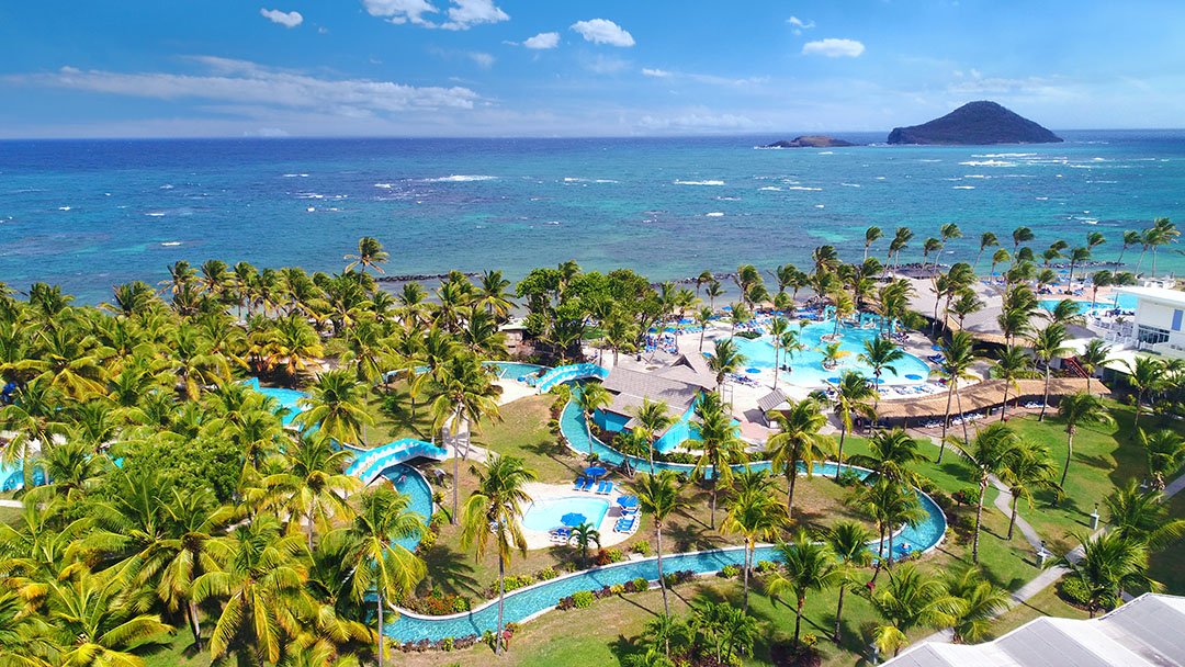 Aerial view of a tropical resort with multiple swimming pools surrounded by palm trees, near the ocean with a small island in the background under a partly cloudy sky.