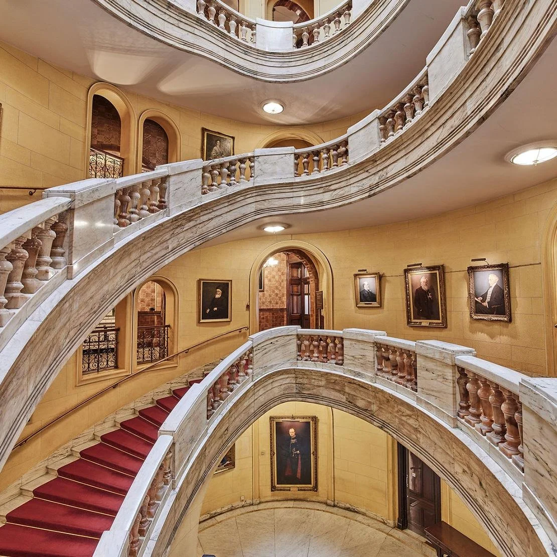 Interior view of a grand staircase with marble balustrades and red carpeted steps, adorned with framed portraits on yellow-painted walls.